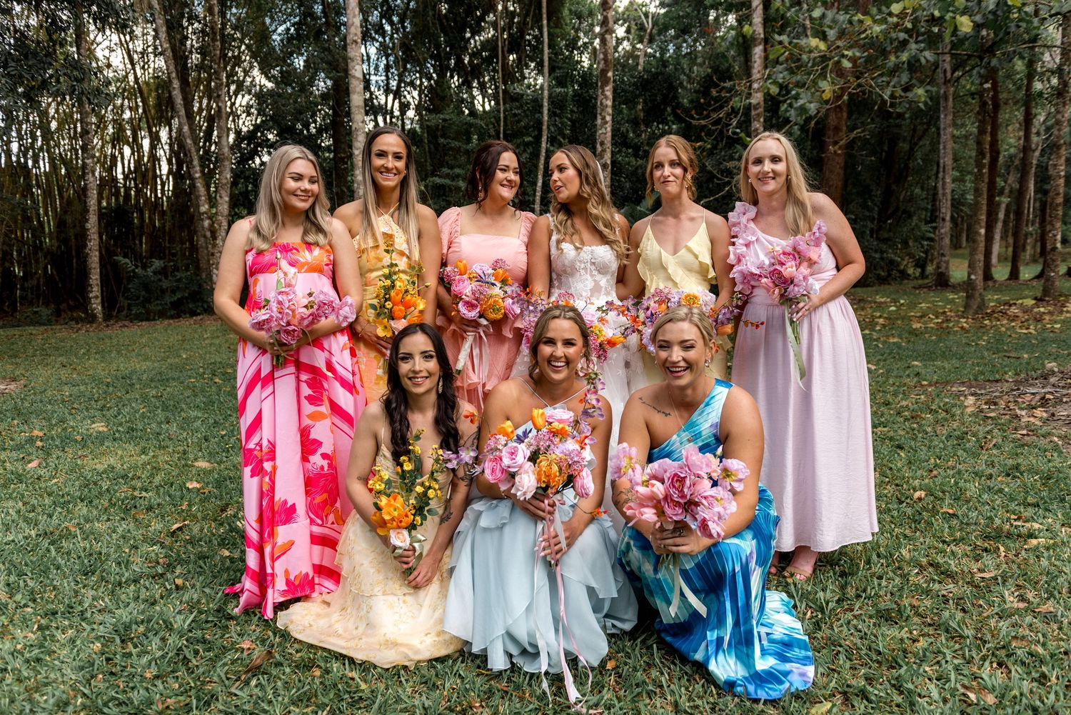 Group of bridesmaids in multicolored dresses posing with bouquets at outdoor woodland wedding.
