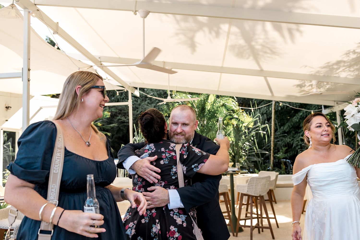 Wedding guests laugh and embrace during outdoor reception celebration under white tent canopy.