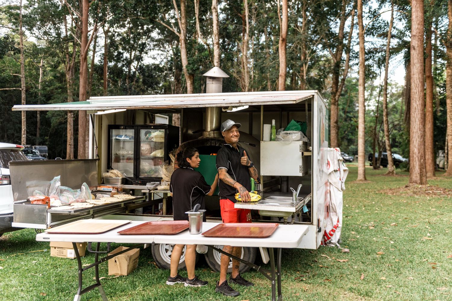 Food truck vendors preparing meals outdoors surrounded by tall eucalyptus trees at outdoor event.