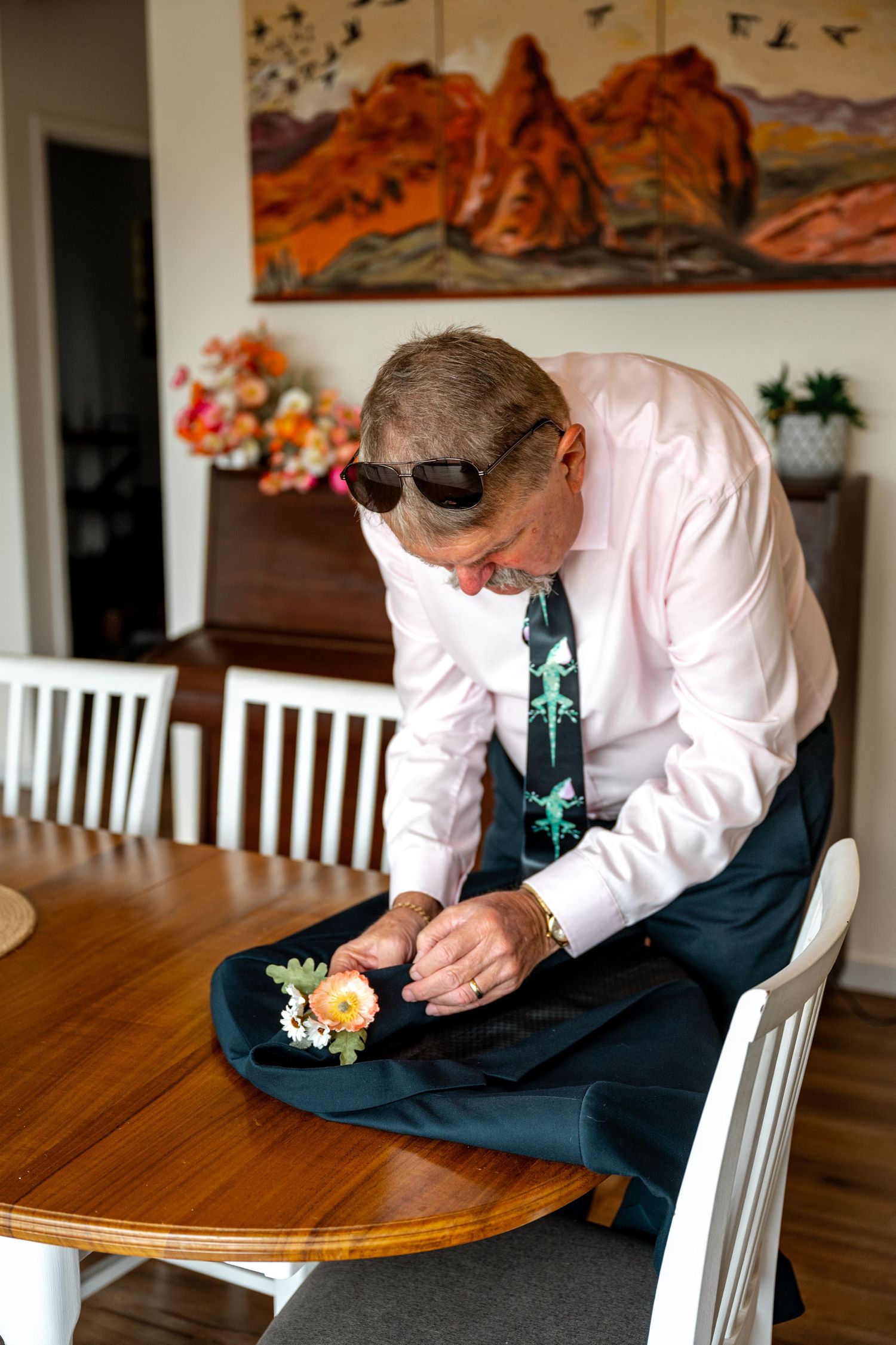 Older man in pink shirt and patterned tie pinning boutonniere onto jacket at wooden dining table.