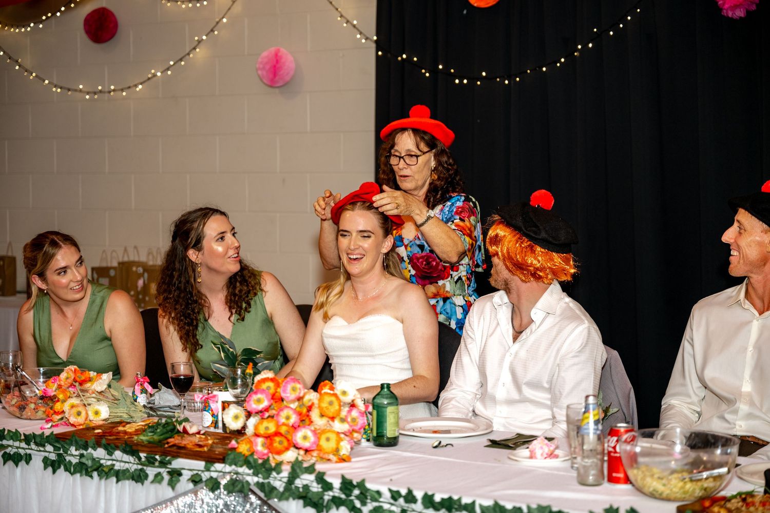 Bride and wedding party laugh at the head table as a guest places a red hat on the bride.