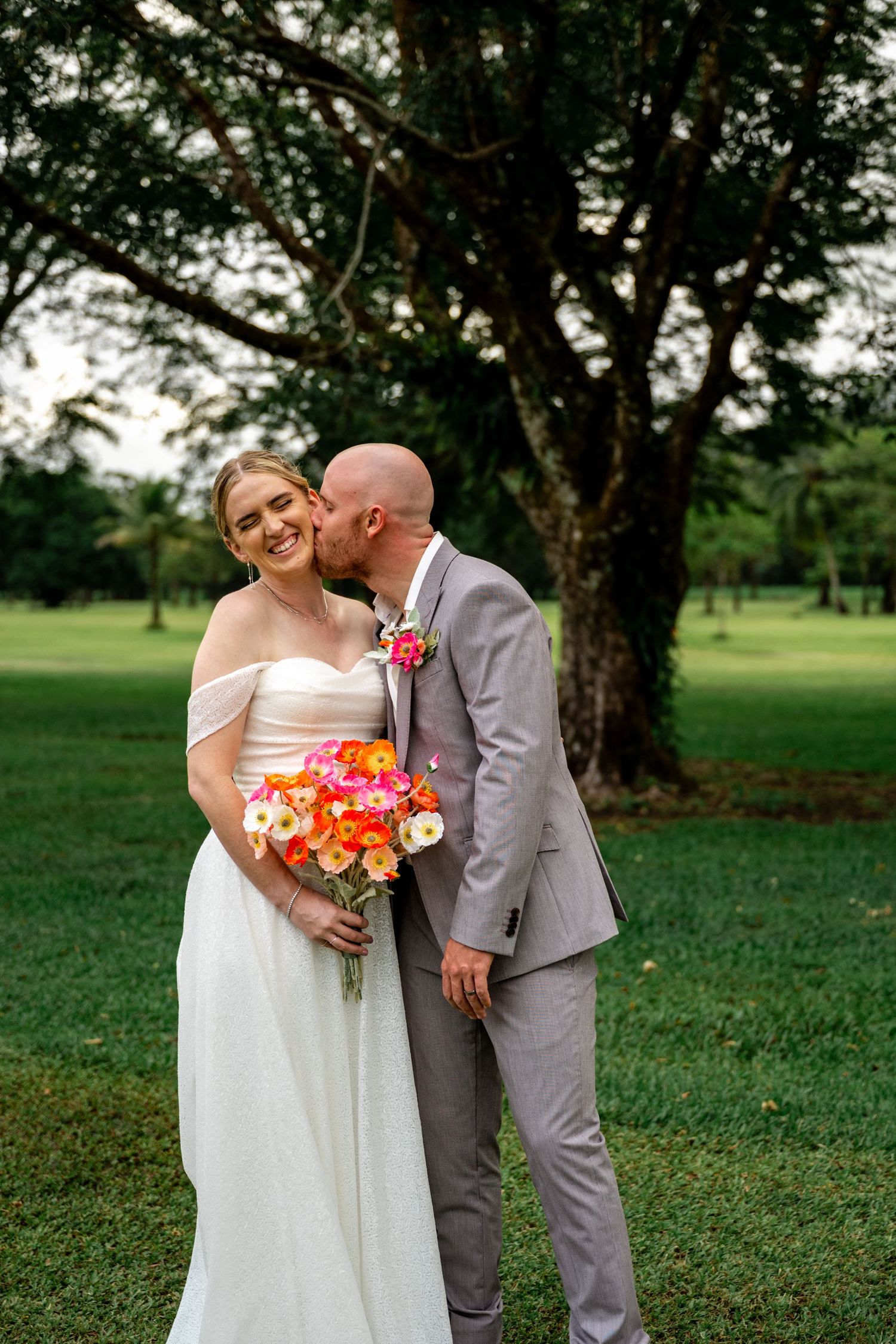 Groom in grey suit kissing bride's cheek as she smiles holding vibrant orange and pink bouquet outdoors.