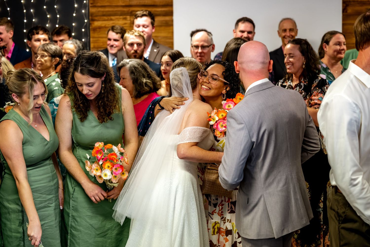 Bride embraces guest while groom smiles during joyful post-ceremony celebrations indoors.