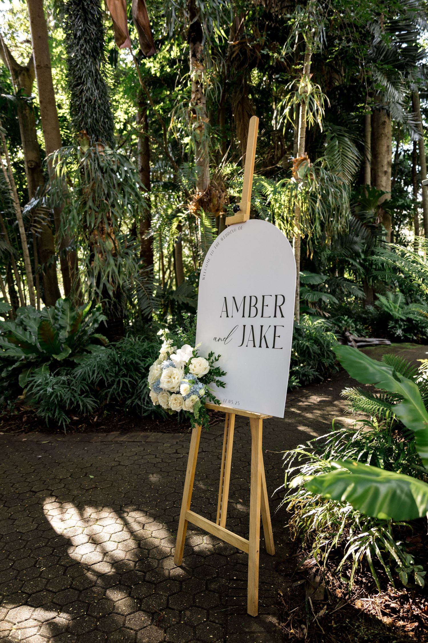 Elegant wedding welcome sign for Amber and Jake on wooden easel adorned with white roses in a garden setting.