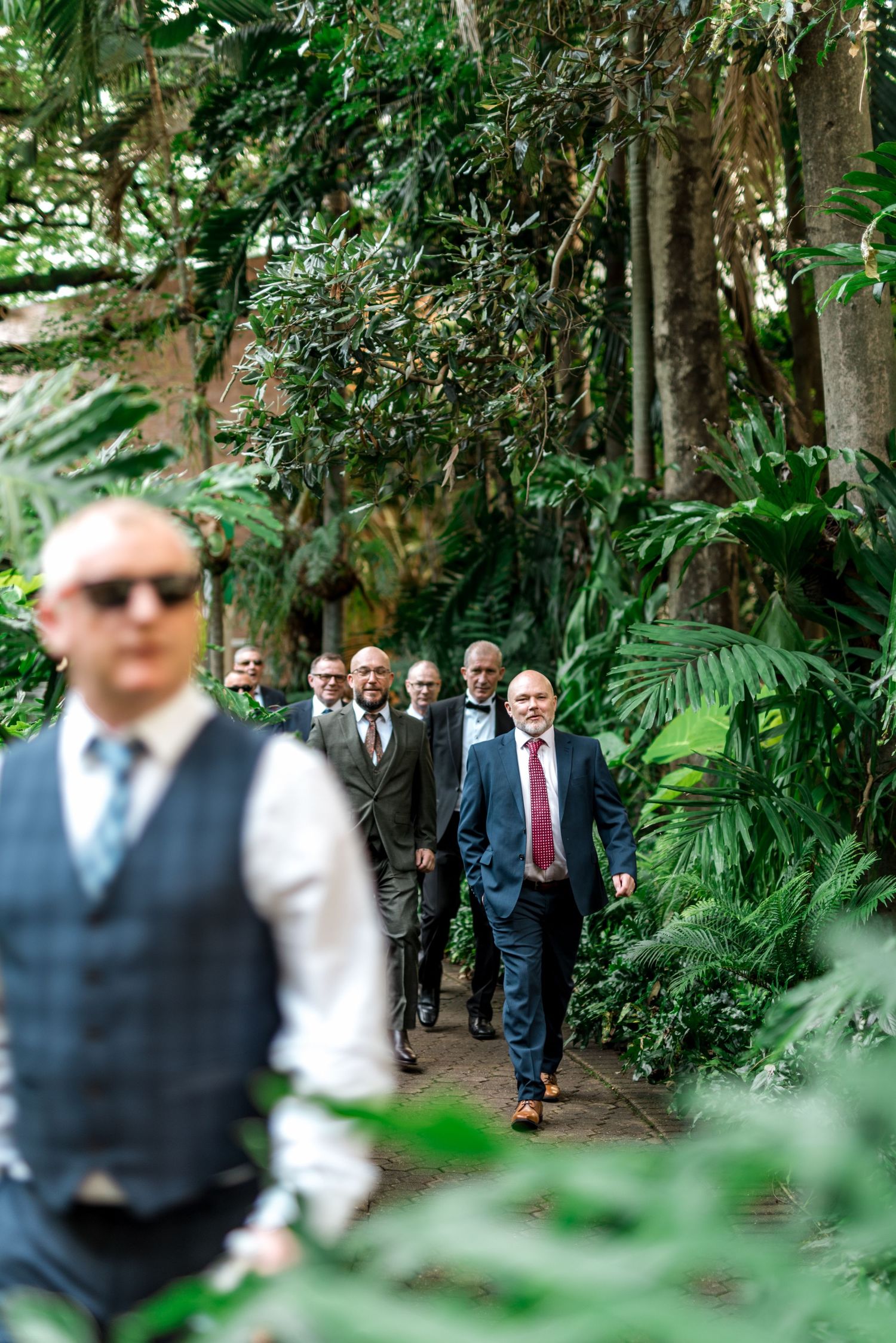 Group of well-dressed men walking along a tropical garden path toward a wedding ceremony venue.