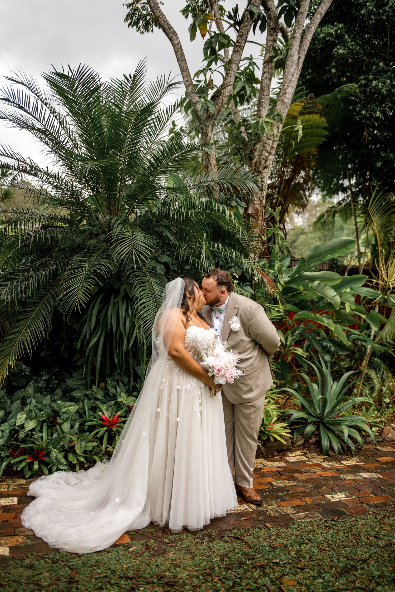 Bride and groom share a kiss surrounded by lush tropical plants and palm trees on their wedding day.
