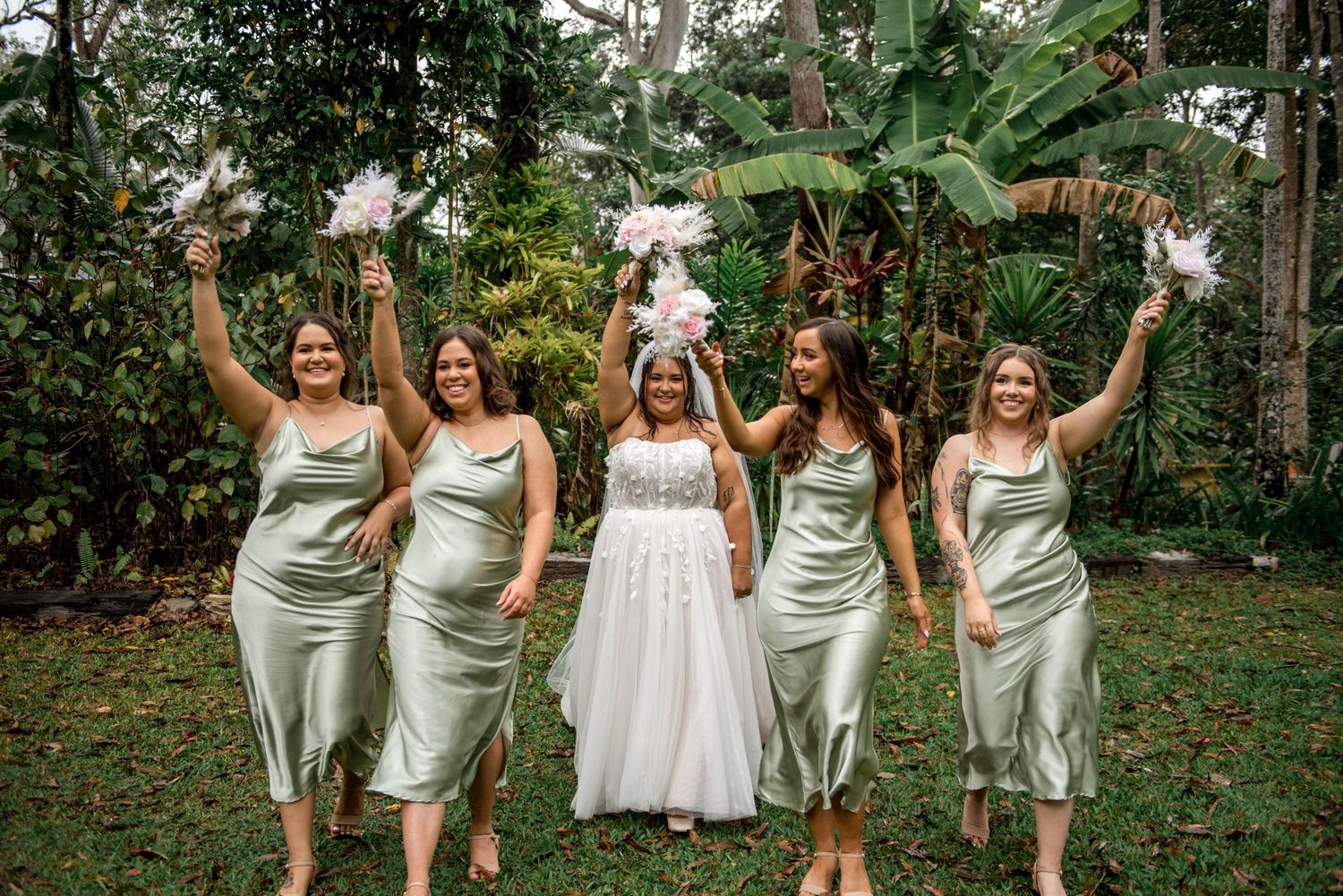 Bride and four bridesmaids in sage green satin dresses raise bouquets joyfully in a lush tropical garden.