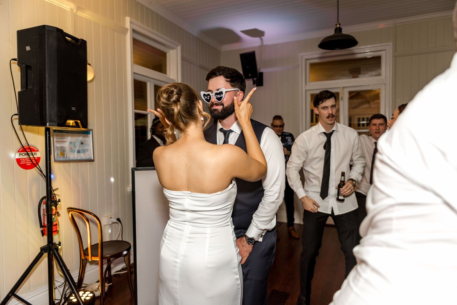 Bride placing heart-shaped sunglasses on groom while dancing at wedding reception.
