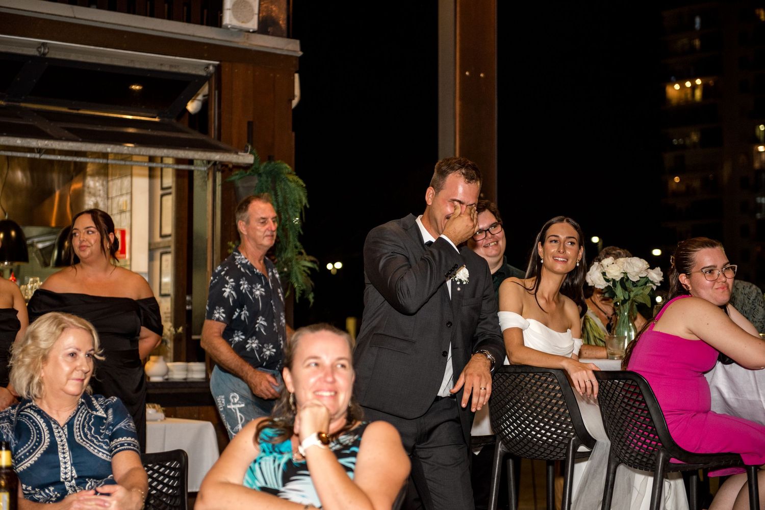 Bride in white dress and groom in tuxedo embrace among seated wedding guests during outdoor evening reception.