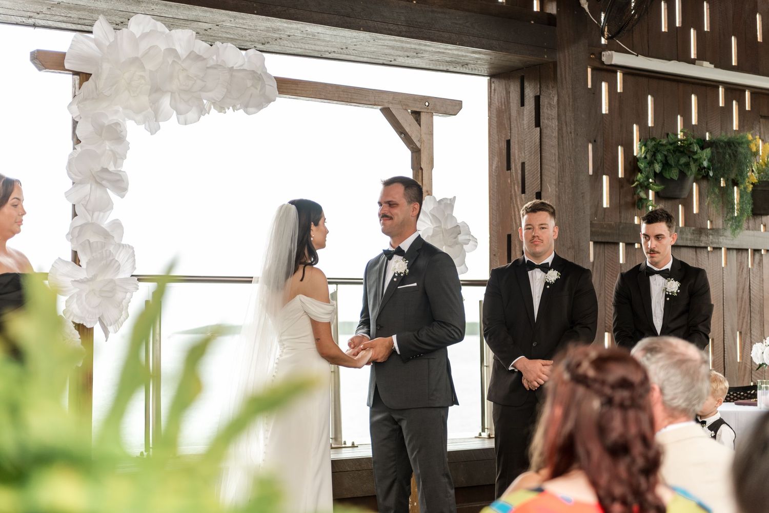 Bride and groom exchange vows under a white floral arch in a rustic wooden wedding venue.