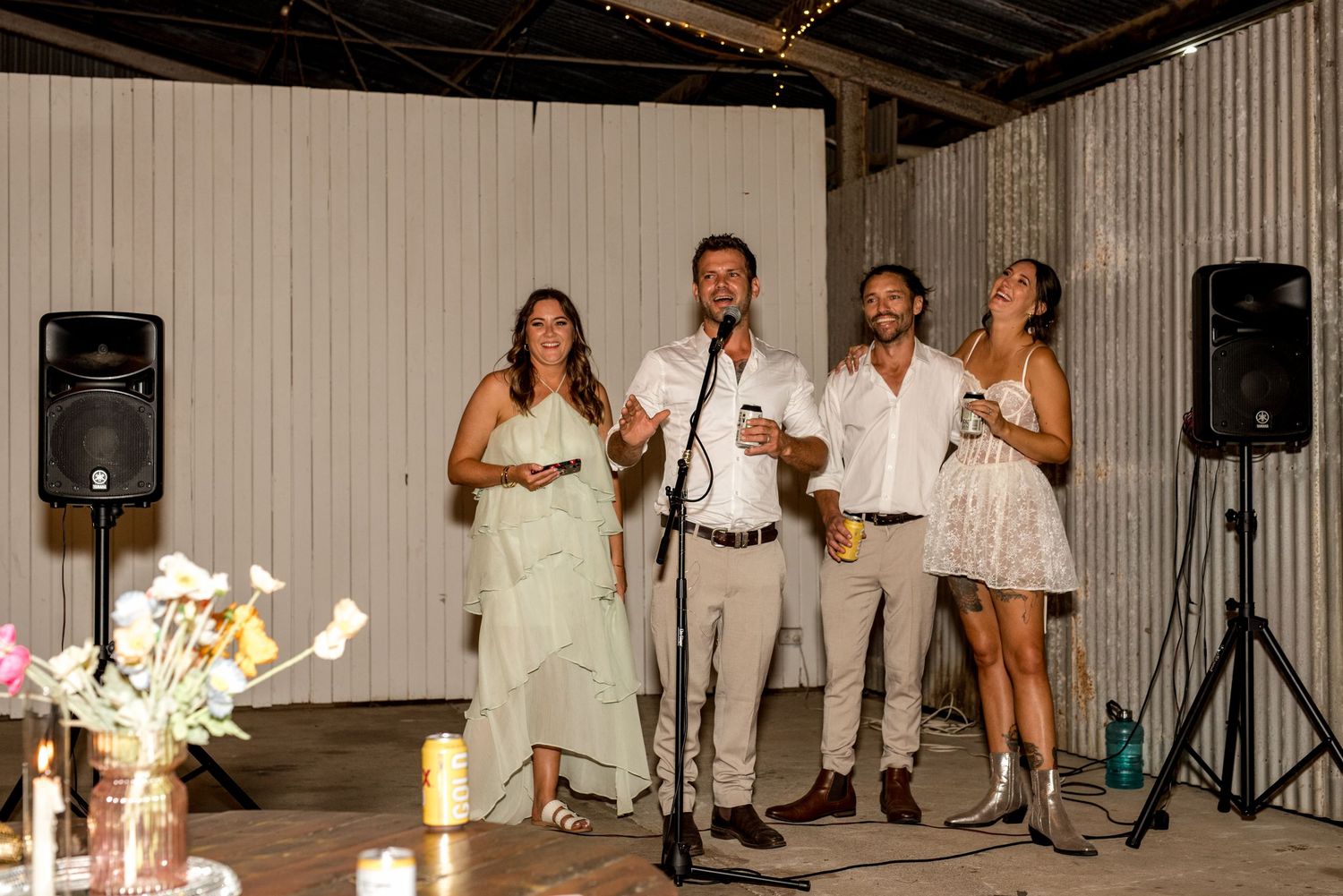 Four guests giving a speech at a wedding reception with speakers and floral centerpieces.