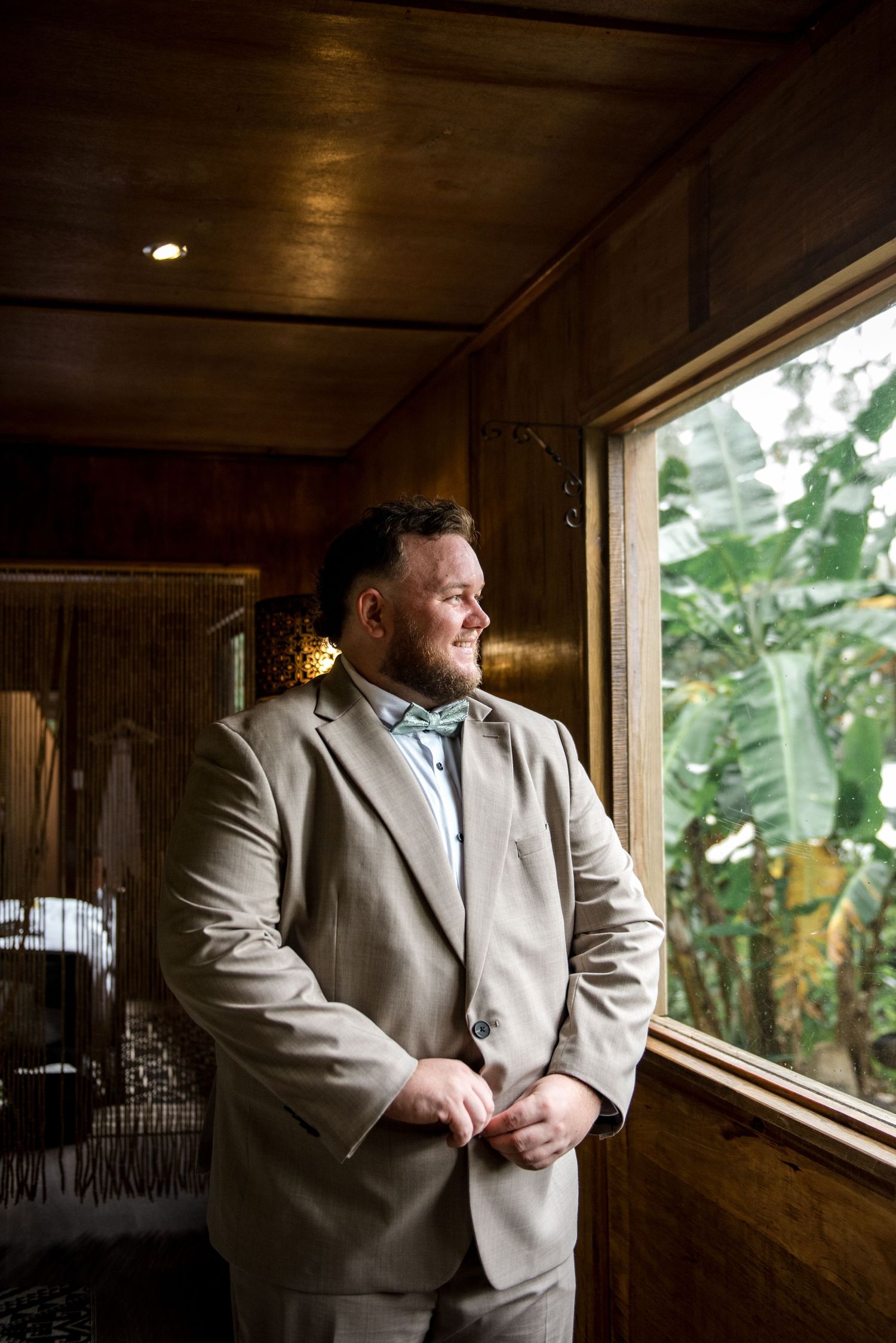 Groom in beige suit with bow tie standing by window in rustic wooden venue smiling.