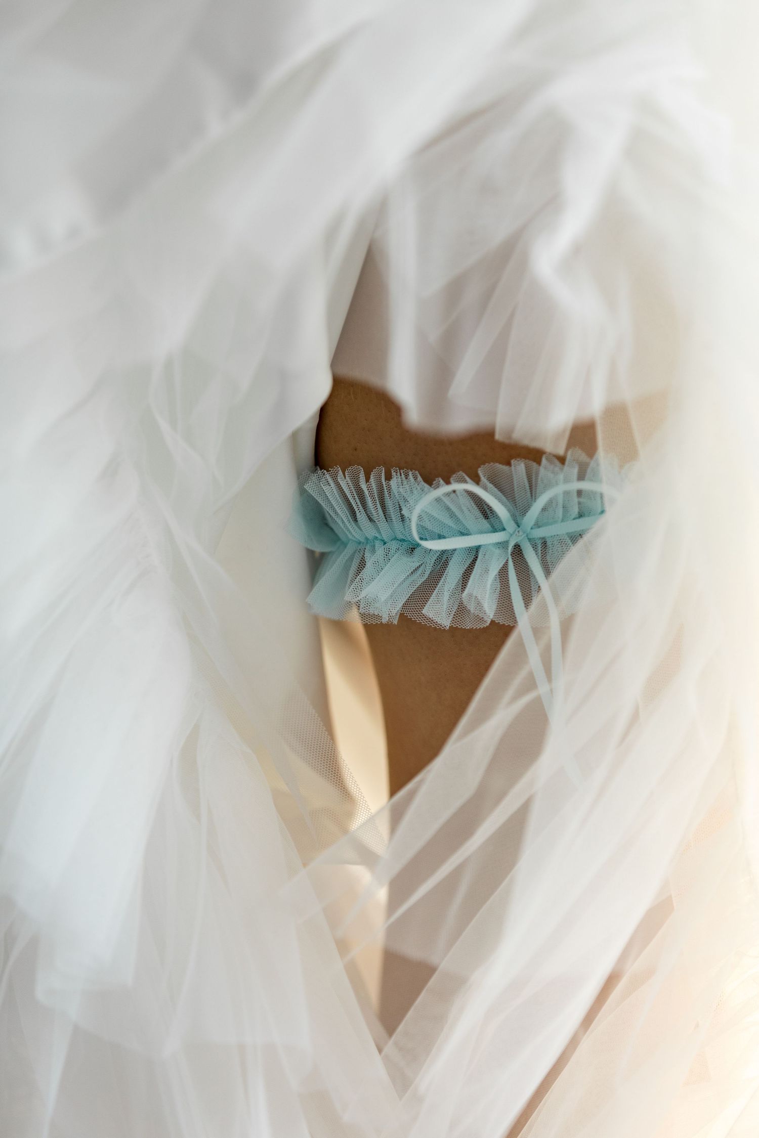 Close-up of bride wearing delicate blue tulle garter beneath white wedding dress.