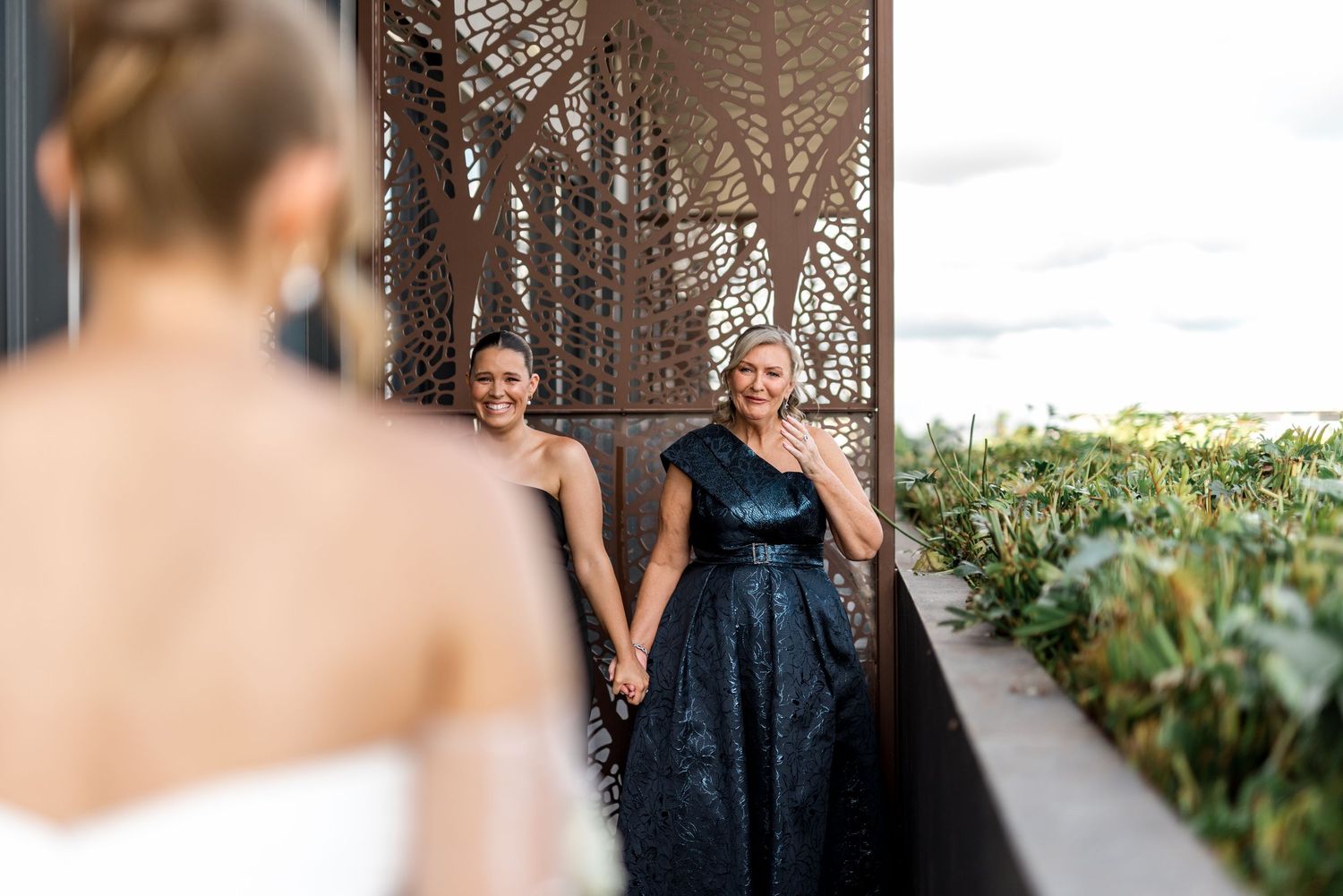 Two guests in black and navy formal gowns holding hands as bride approaches on outdoor terrace.