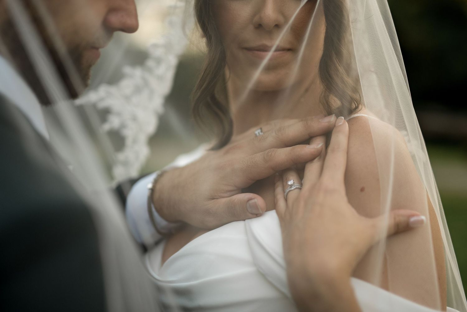 Close-up of groom tenderly touching bride's shoulder through sheer wedding veil, diamond engagement ring visible.
