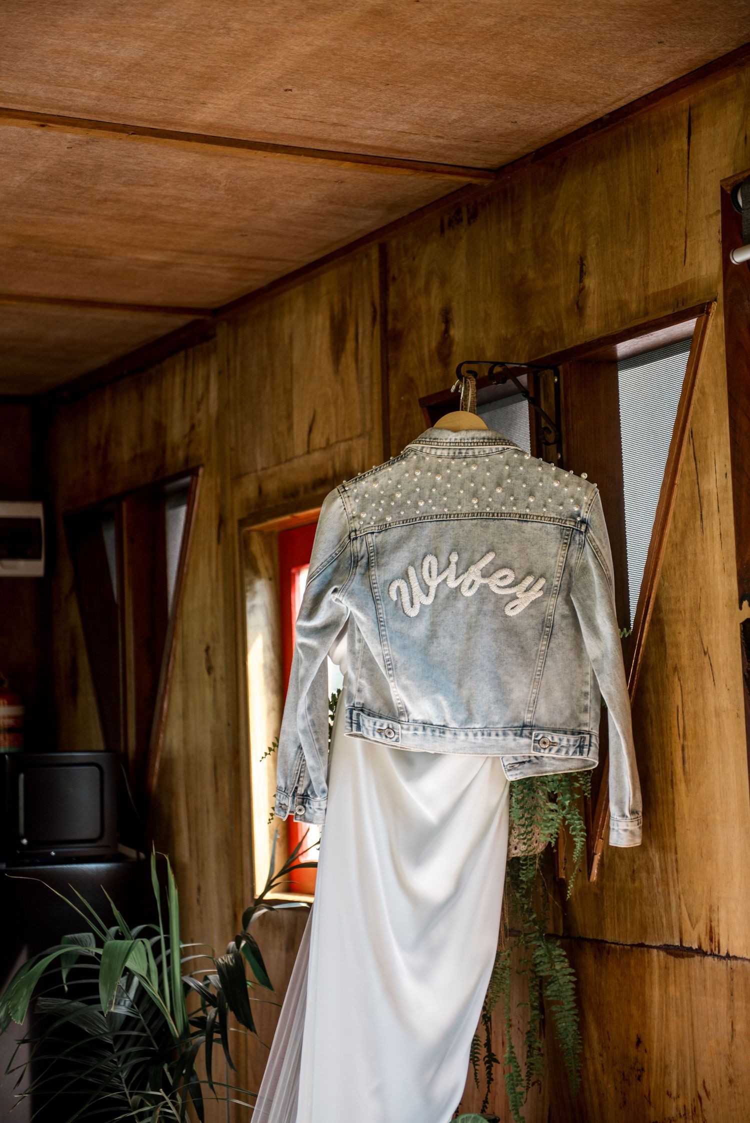 Pearl-embellished denim jacket with Wifey script hanging in a rustic wooden cabin.