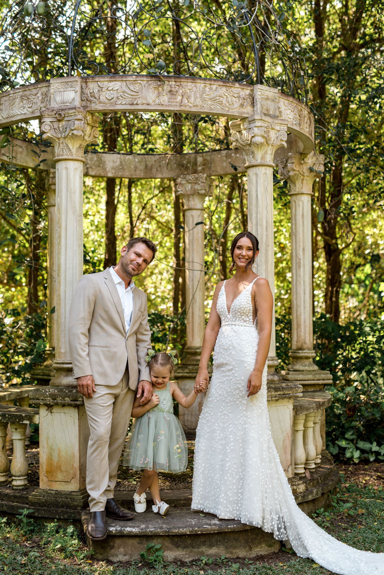 Wedding family portrait with bride, groom, and daughter standing before elegant stone rotunda in lush garden.