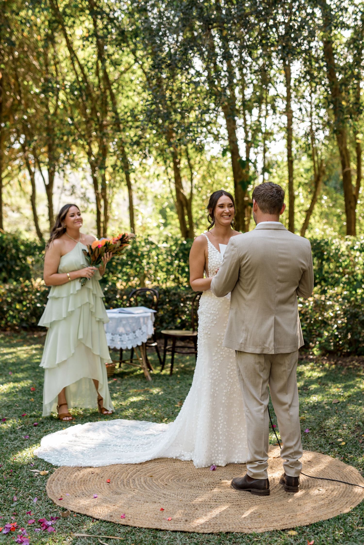 Bride and groom exchange vows outdoors in a lush garden ceremony with bridesmaid holding colorful bouquet.