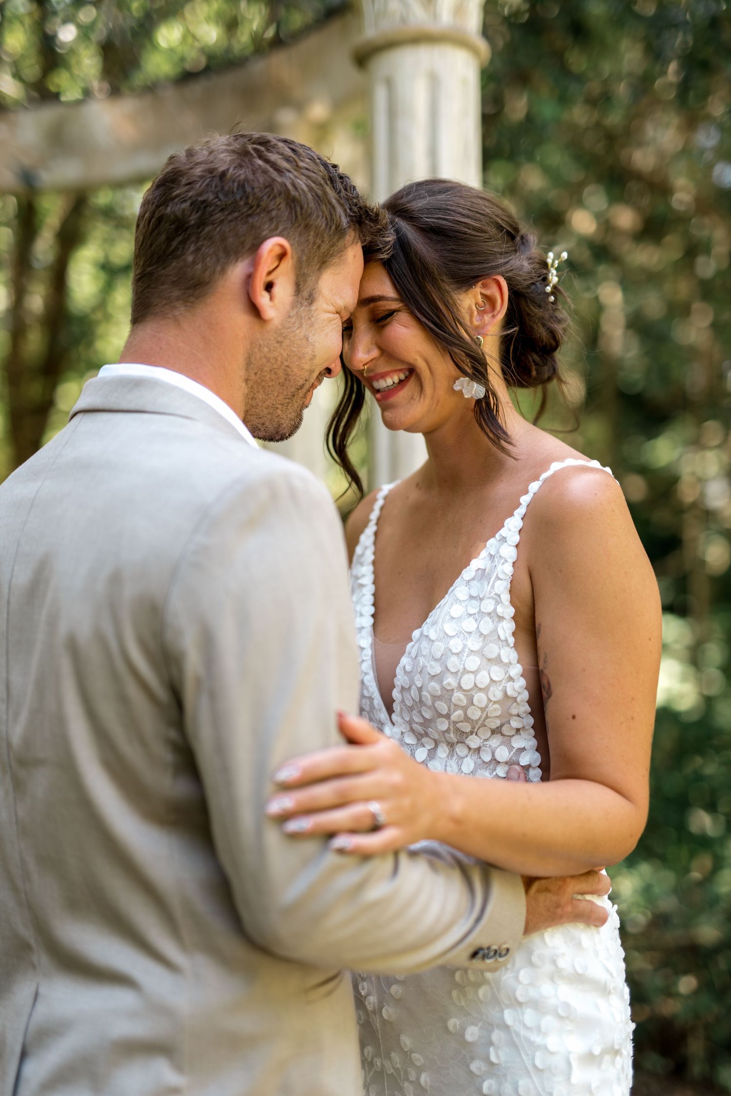 Smiling bride in floral applique gown shares tender forehead touch with groom near stone garden gazebo.