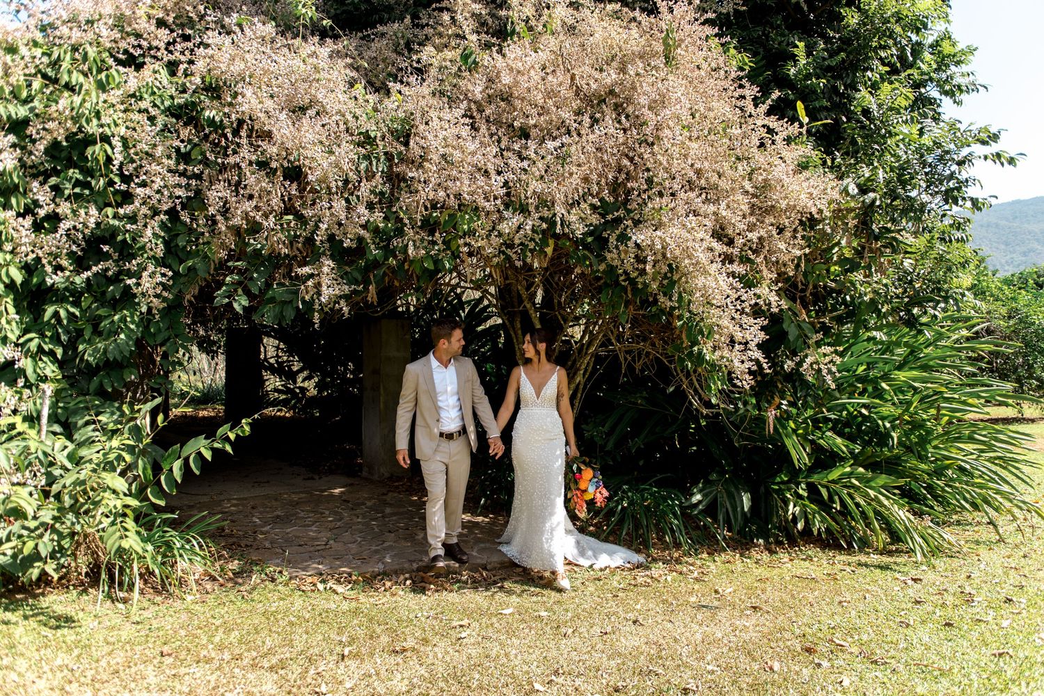 Newlyweds walk hand in hand beneath a flowering tree with colorful bridal bouquet outdoors.