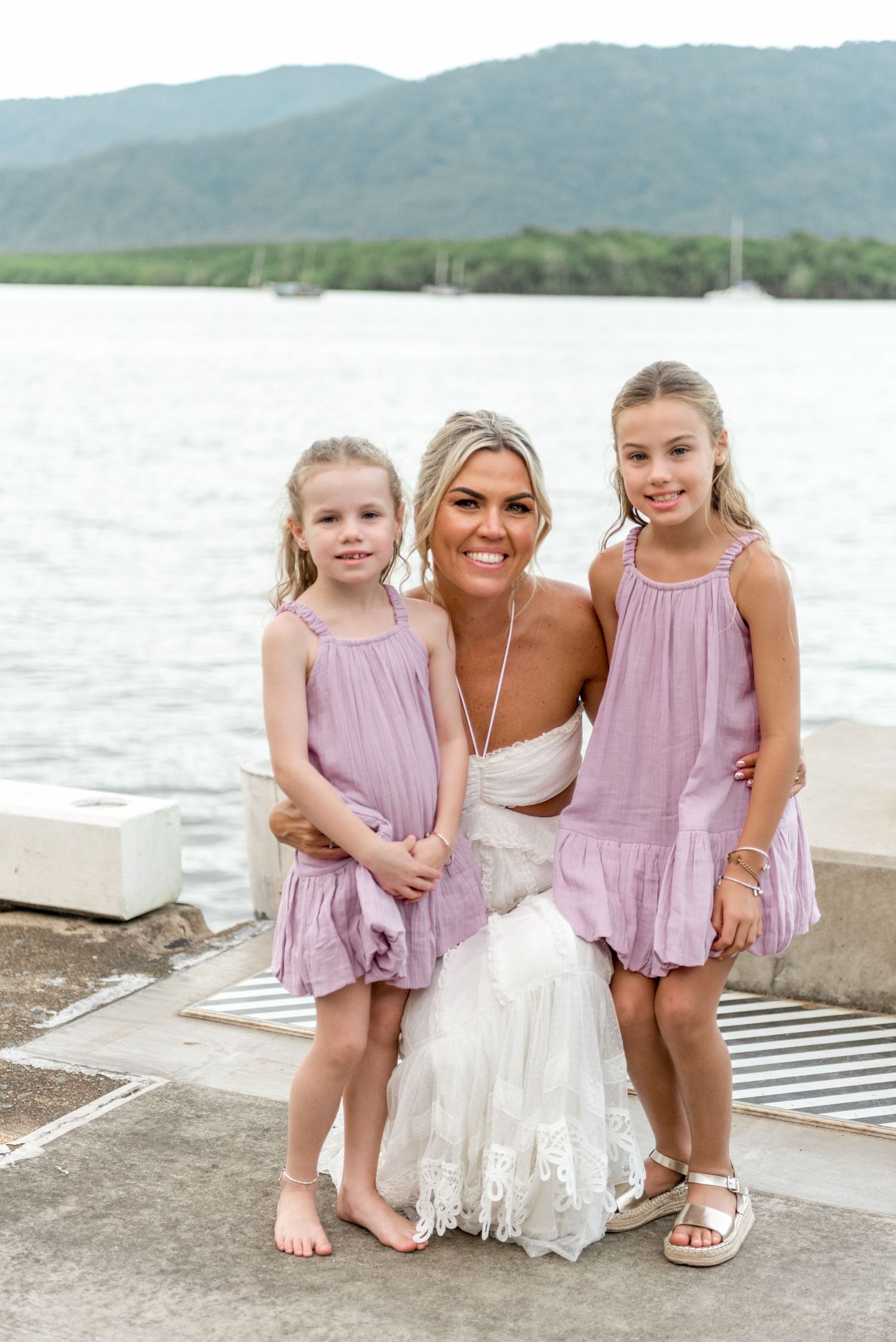 Smiling bride posing with two young flower girls in matching lavender dresses by waterfront.