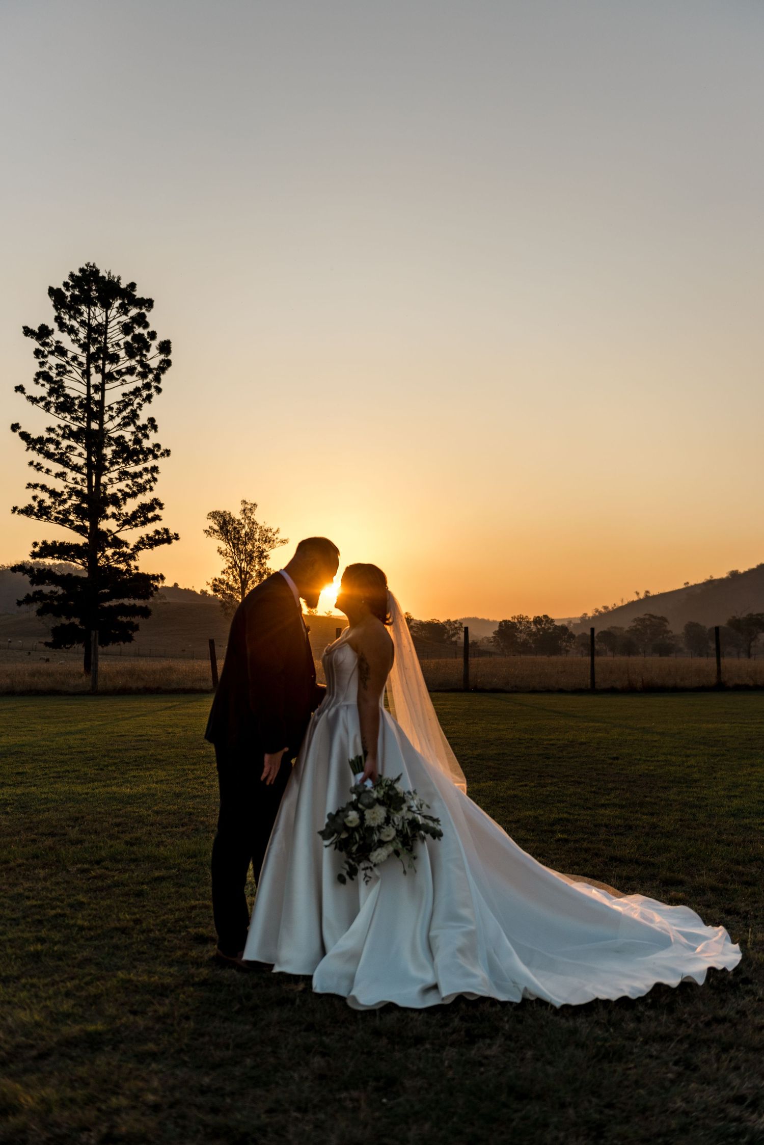 Bride and groom silhouetted against vibrant golden sunset with tall pine tree in rural landscape.