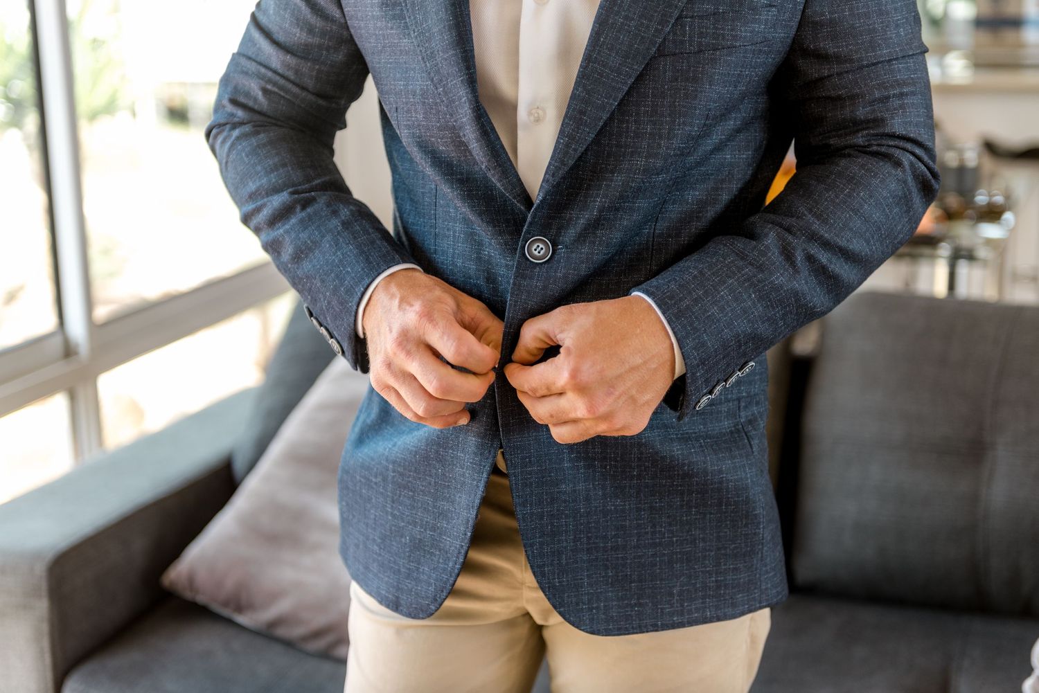 Groom buttoning navy blue checkered blazer over beige trousers during wedding day preparations.