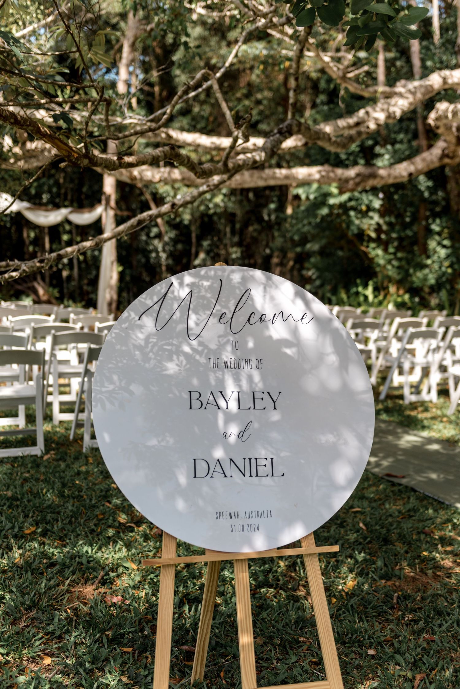 Round welcome sign for Bayley and Daniels wedding displayed on a wooden easel outdoors.