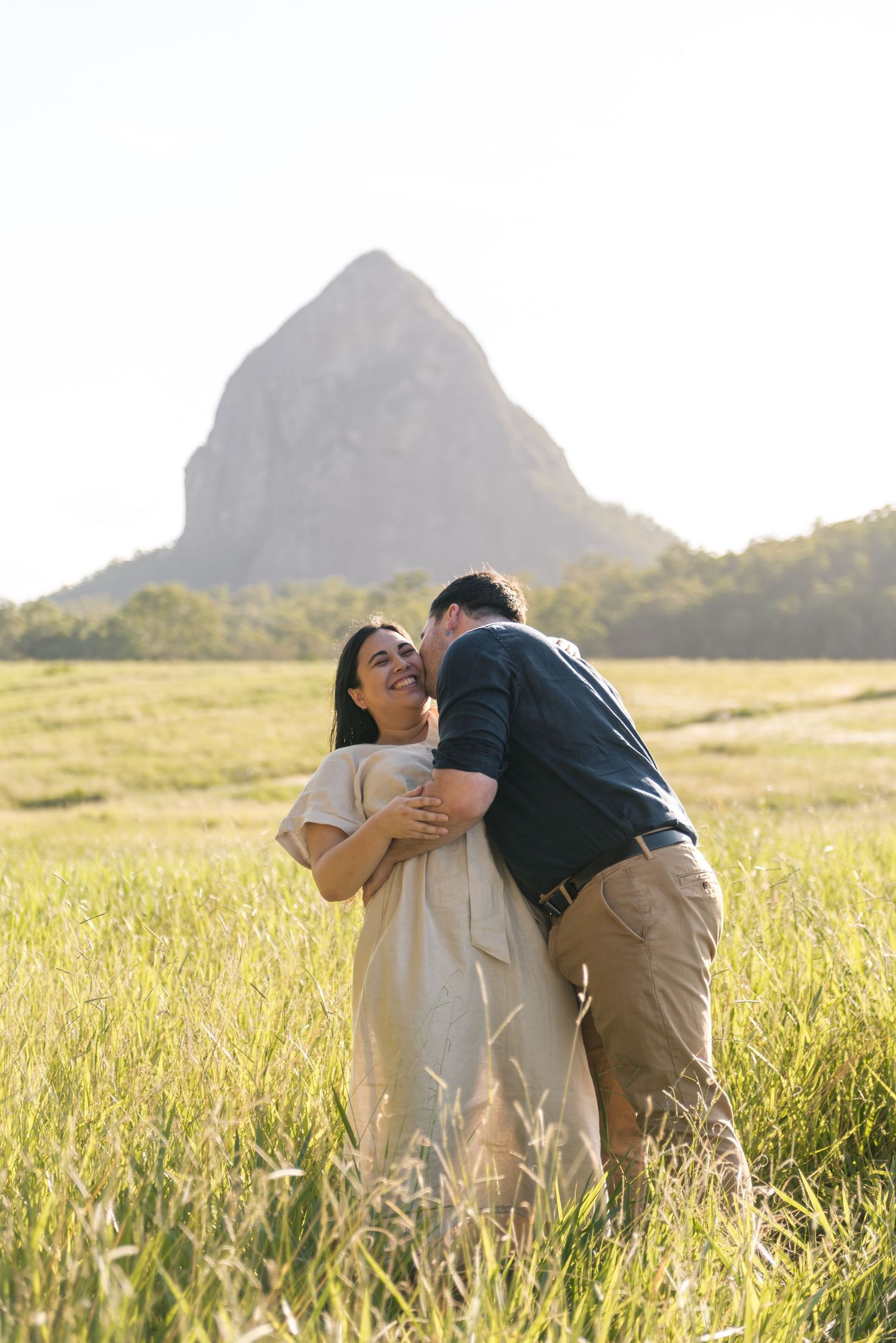 Man kissing woman on the cheek as she laughs in a sunlit field with a rocky mountain backdrop.