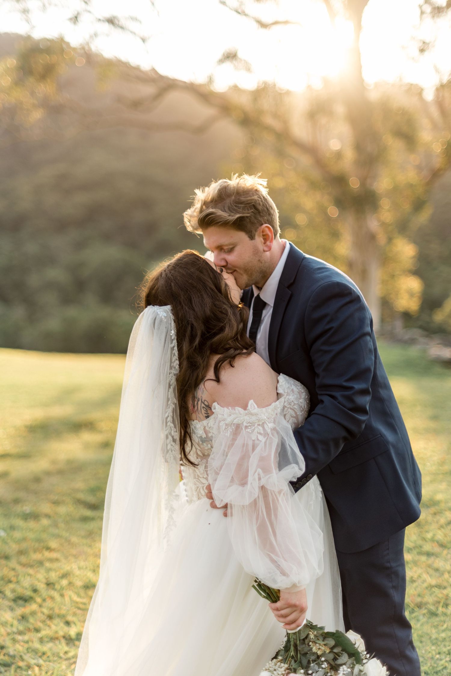 Bride and groom sharing romantic kiss bathed in golden sunset light on green lawn.