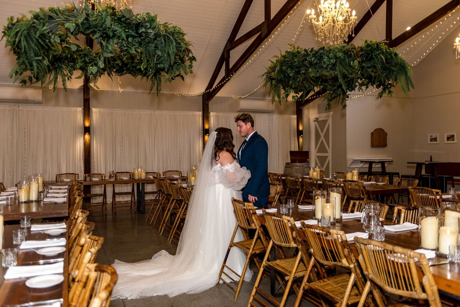 Bride and groom share a romantic moment in an elegant barn wedding reception hall with greenery chandeliers.