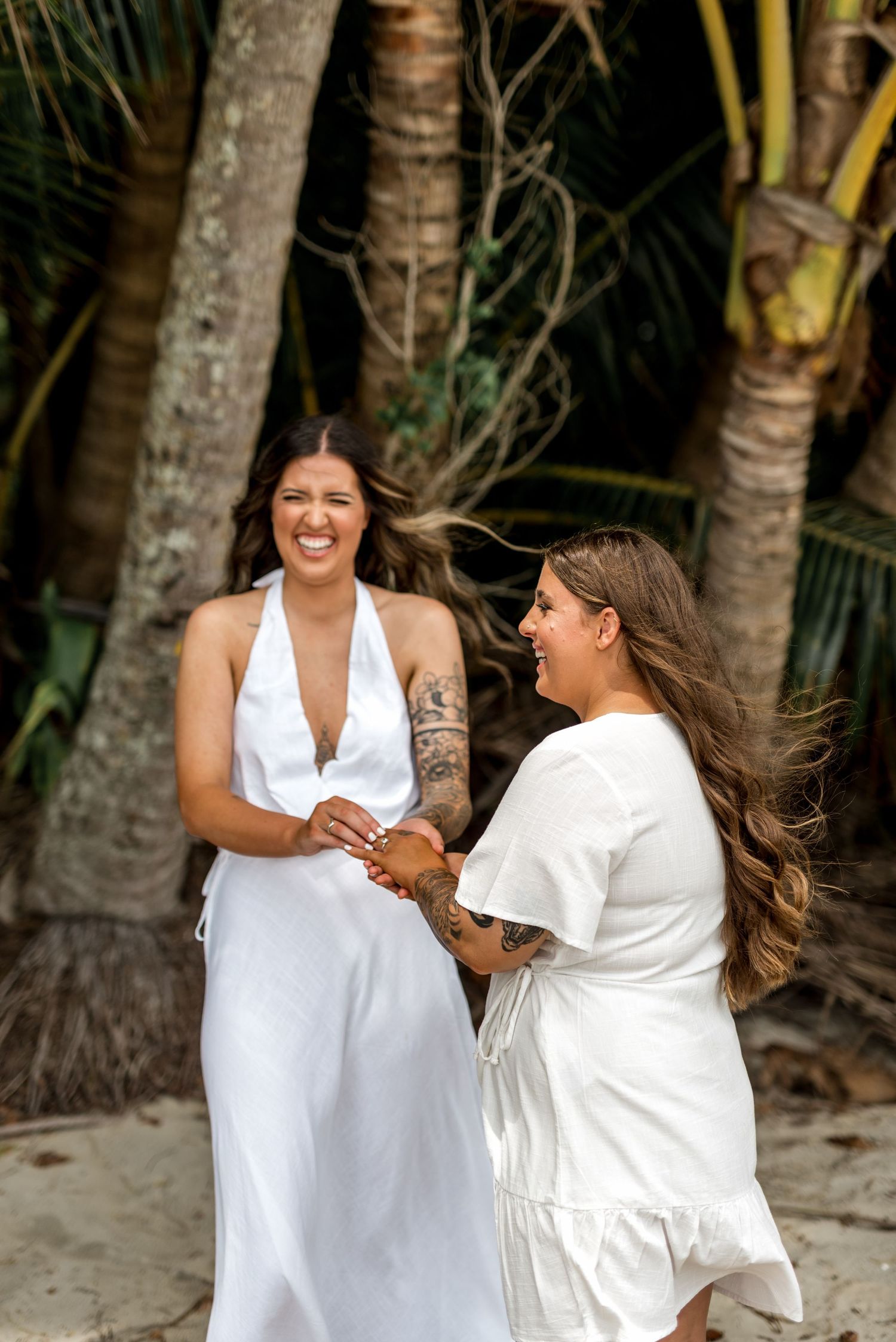 Two brides laughing joyfully while exchanging rings during their tropical beach wedding ceremony.