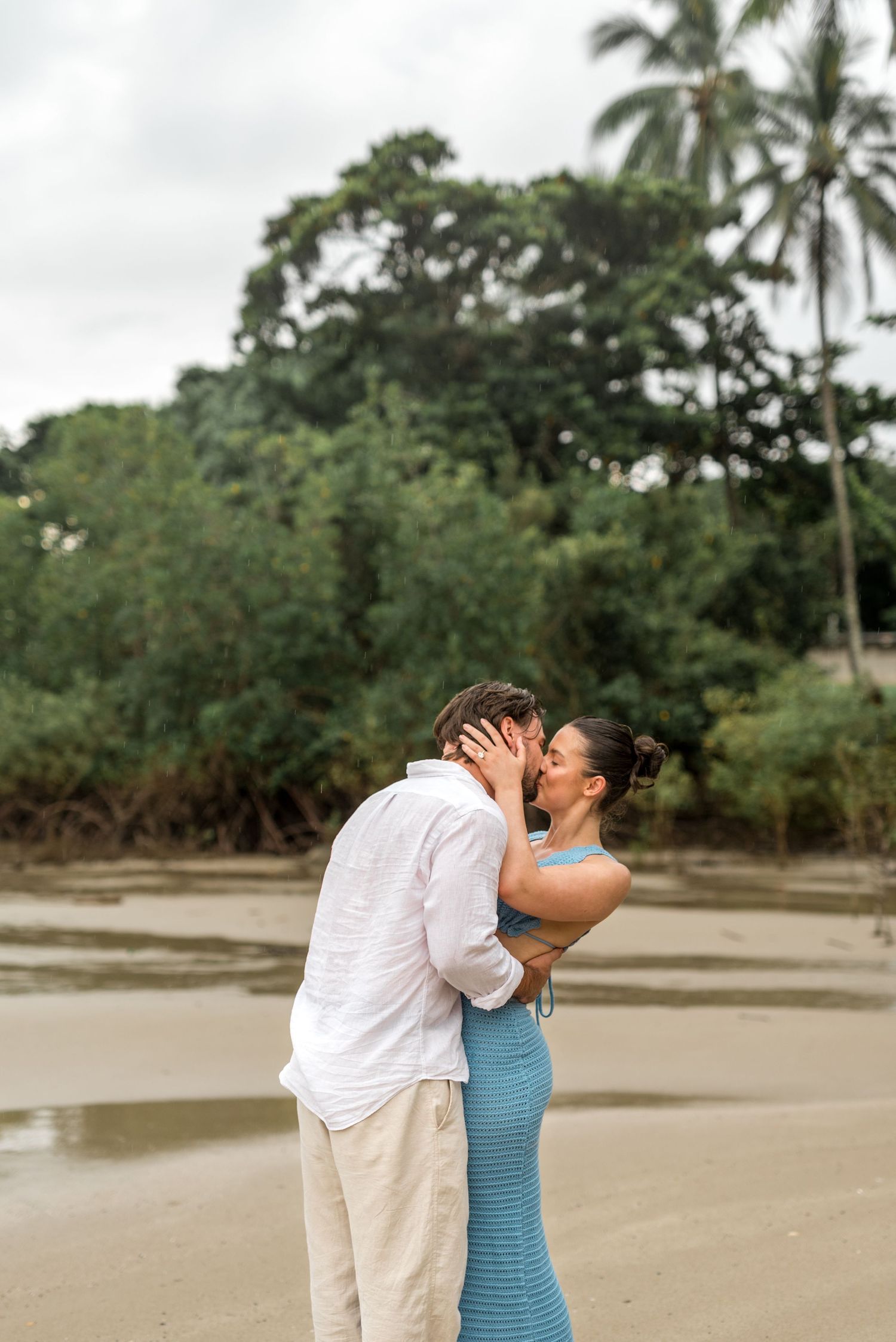 Couple sharing a tender kiss on a sandy beach with lush tropical trees in background.