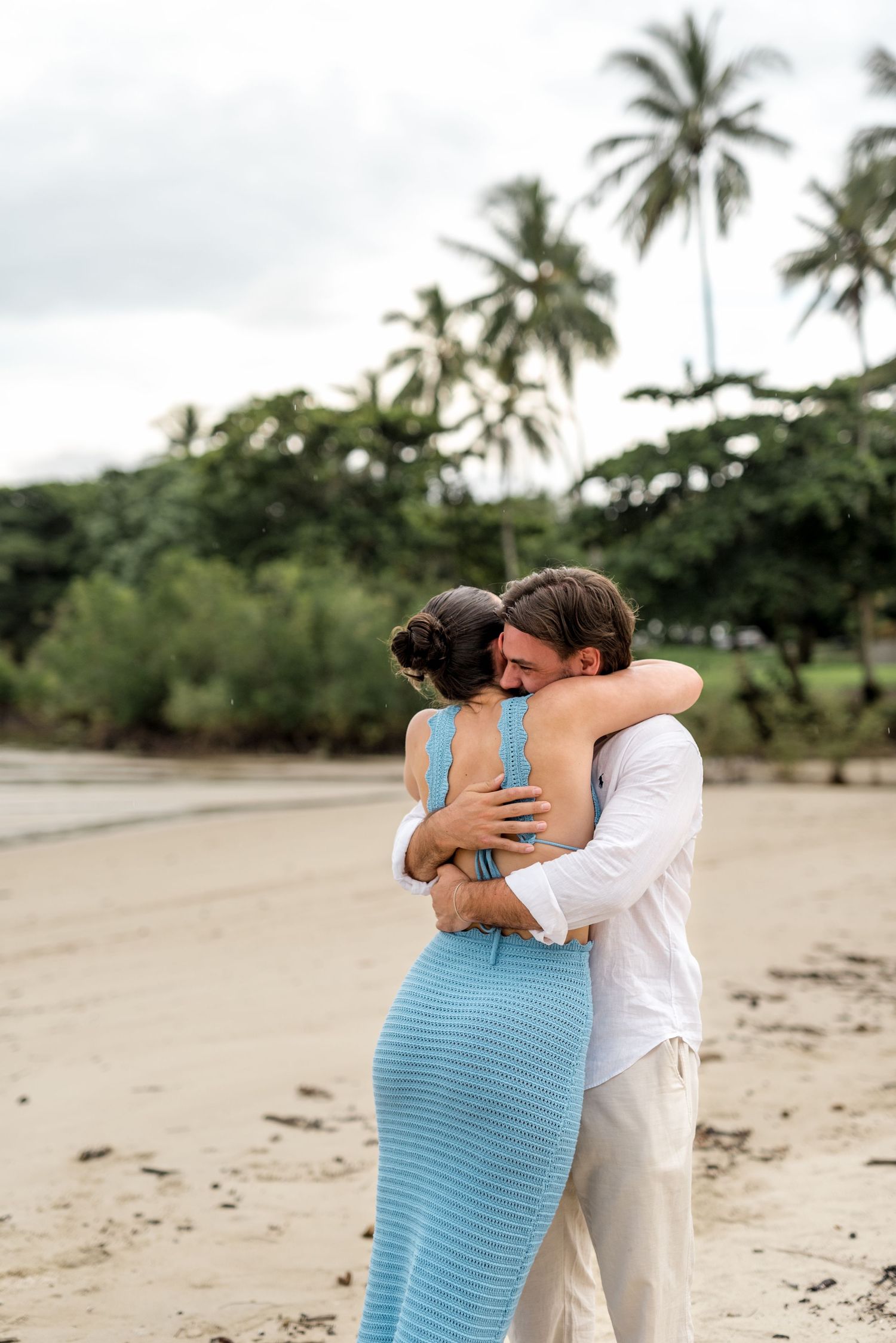 Couple embracing in a tender hug on a tropical beach surrounded by lush palm trees on a cloudy day.