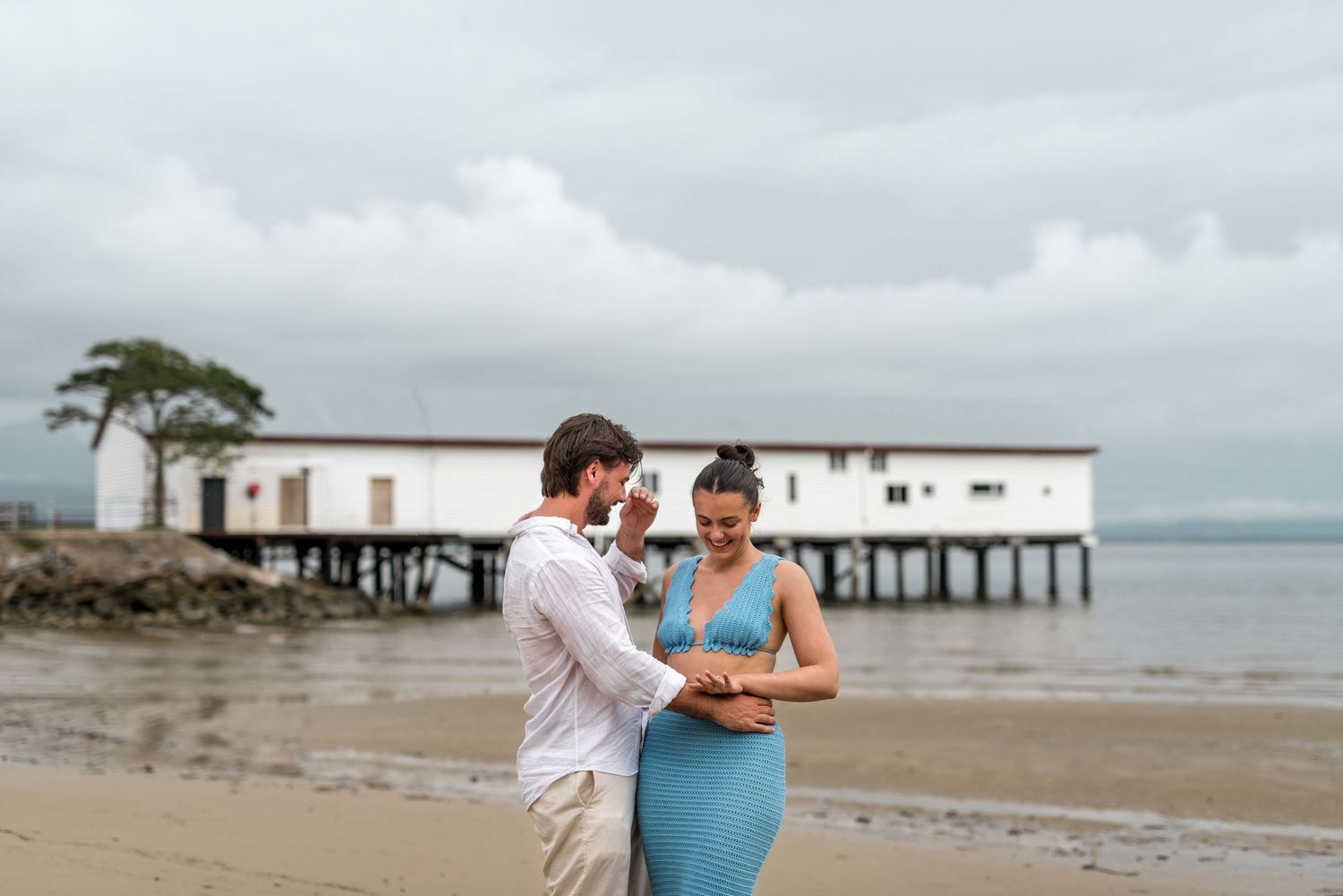Couple looking down together on a sandy beach with a white waterfront building on stilts in the background.