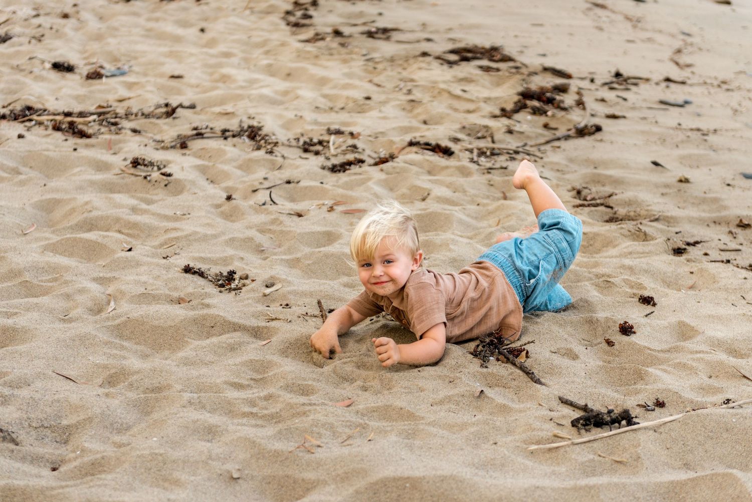 A happy blonde toddler lies on sandy beach smiling at the camera during a family outing.