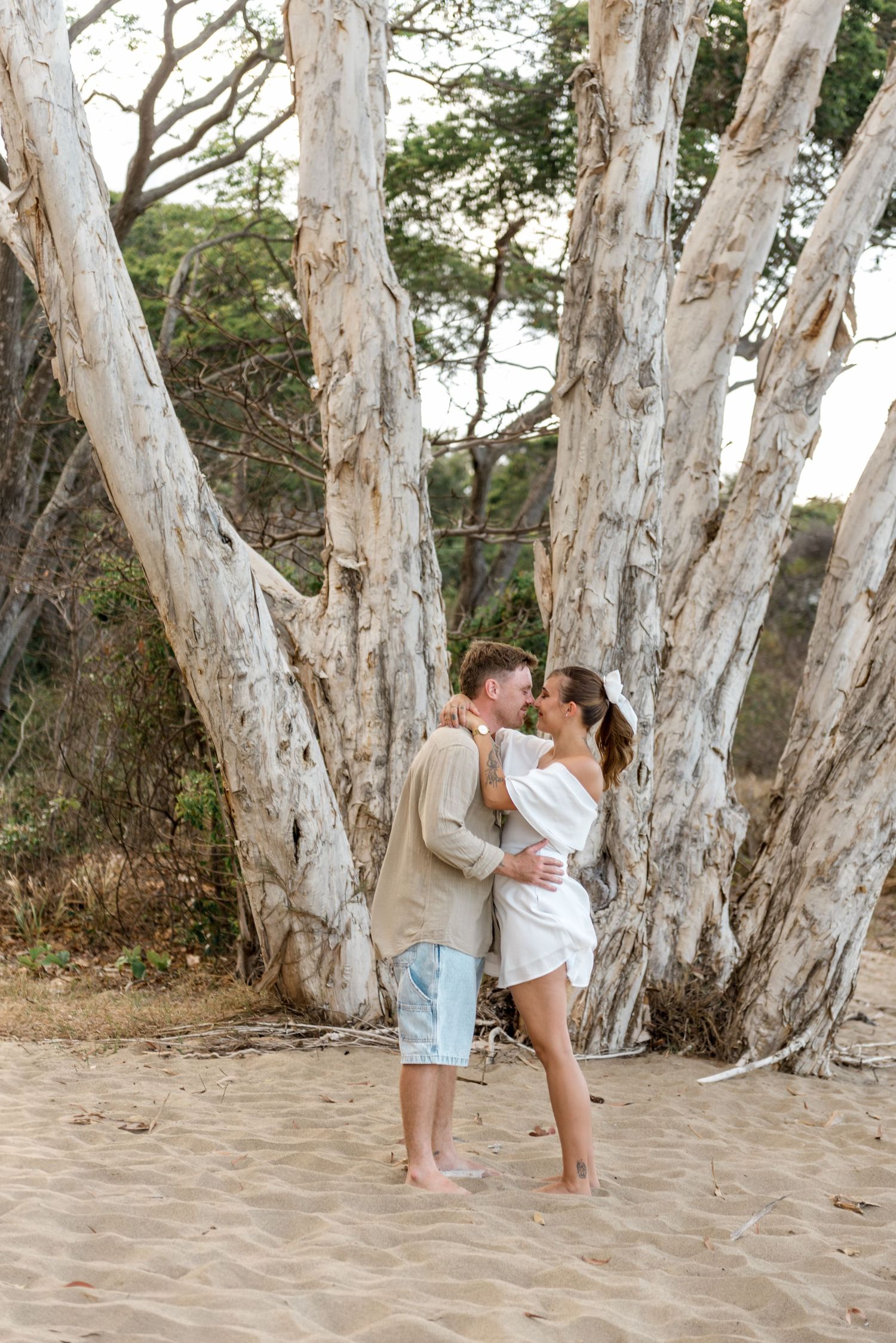 Couple laughing and embracing on sandy beach with striking white paperbark trees in background.
