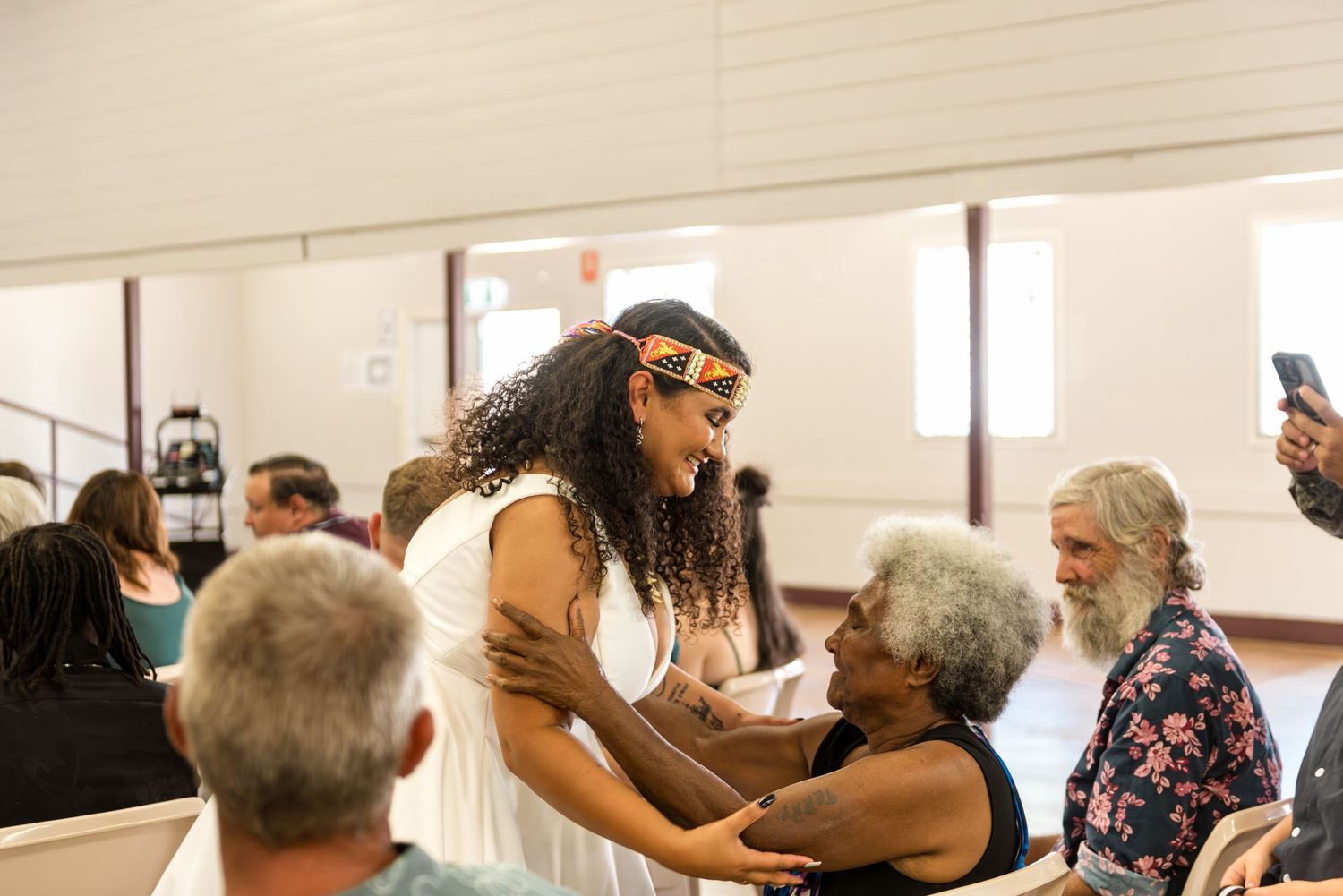 Bride in white dress with cultural headpiece embraces elderly guest warmly after wedding ceremony indoors.