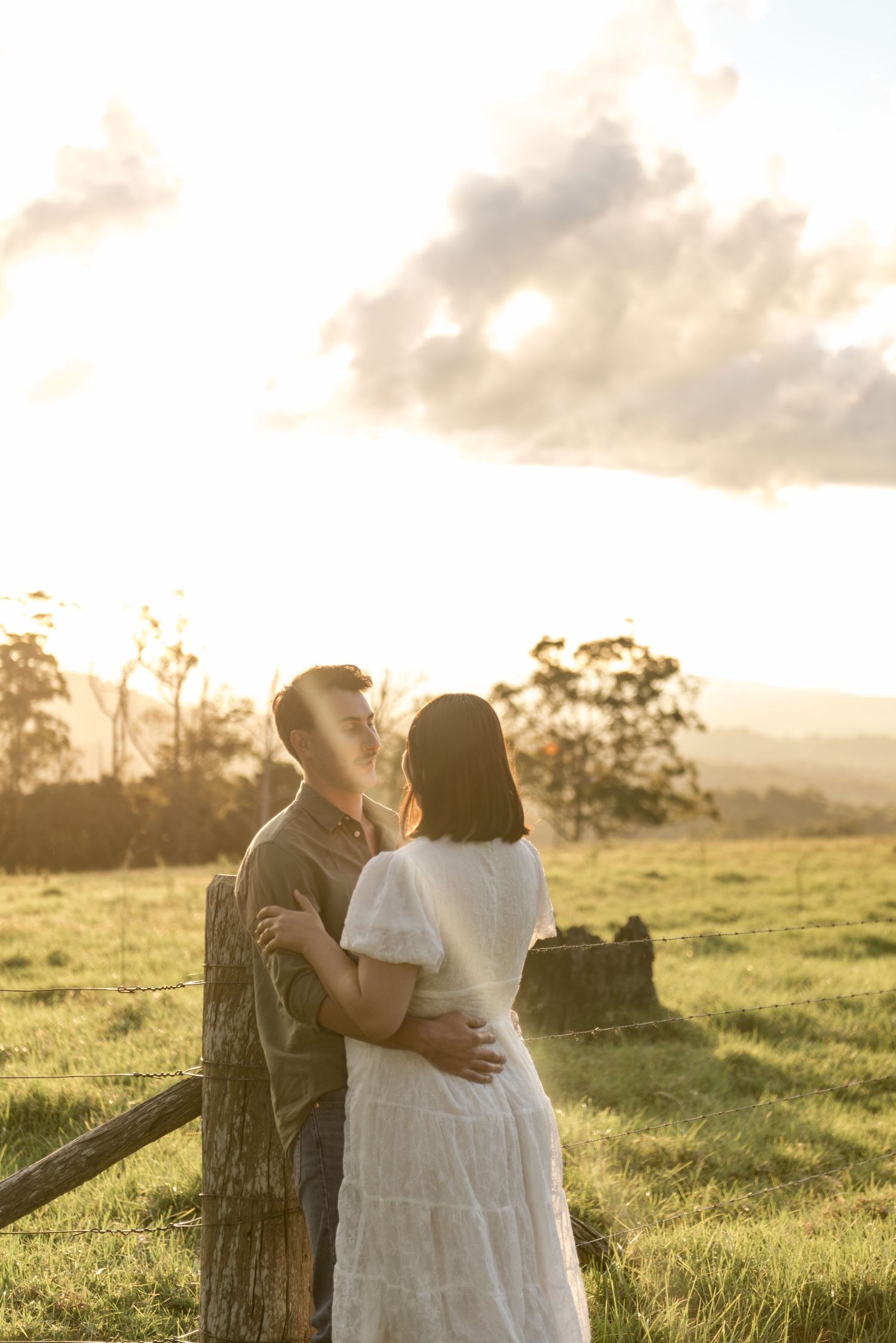 Couple embracing at golden hour by a rustic fence in a lush green field.