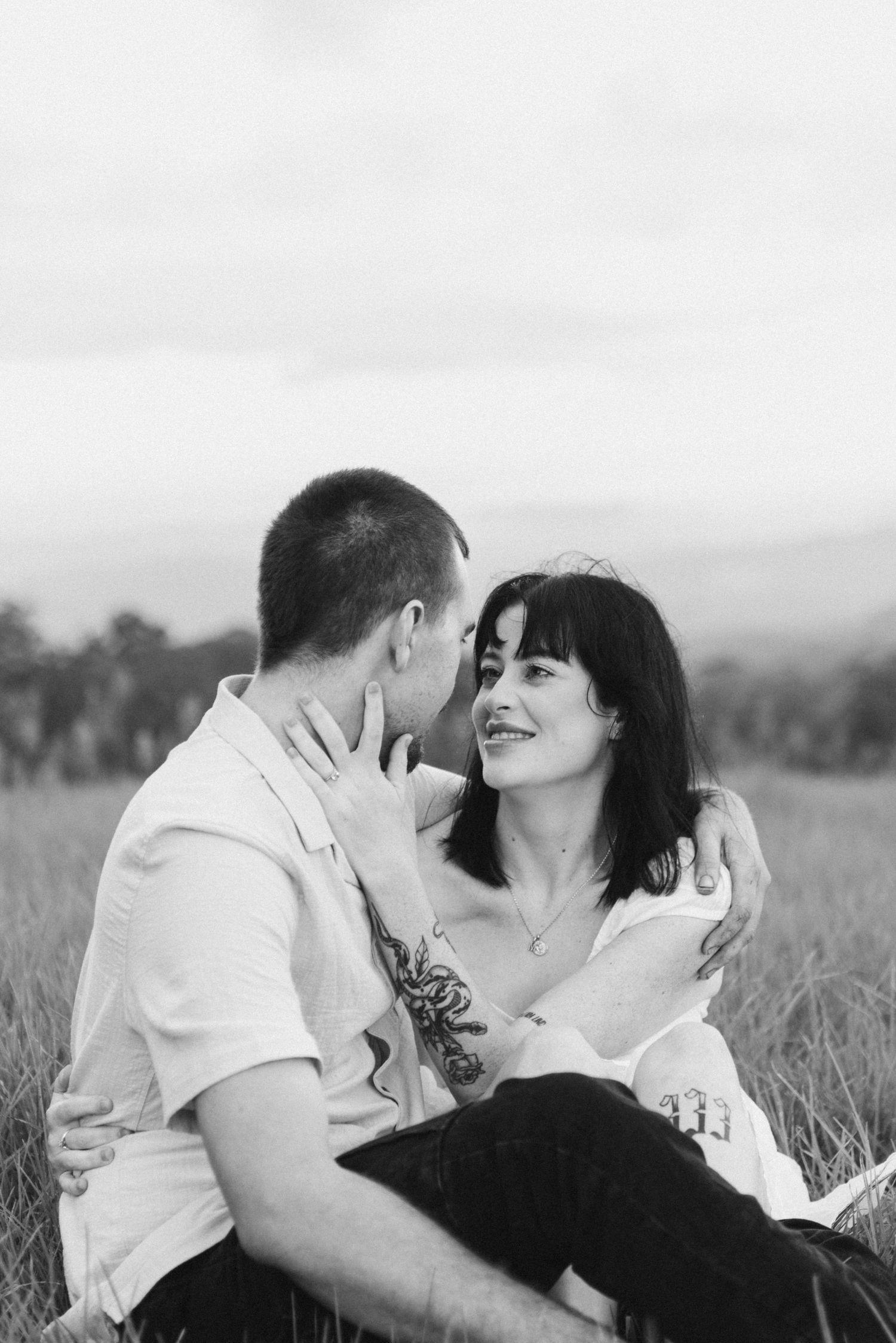 Black and white photo of couple sitting in field, woman smiling at man while embracing tenderly.
