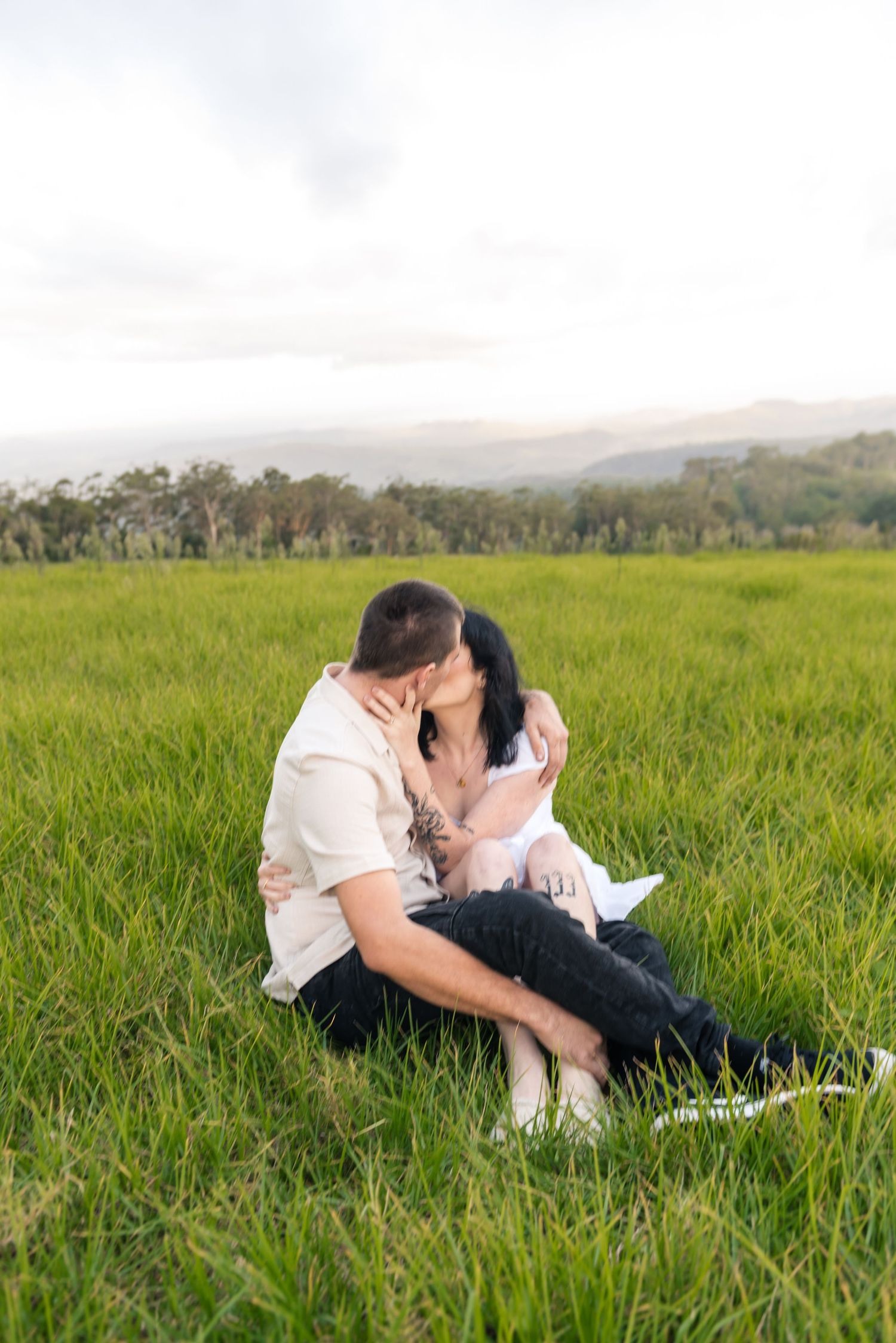 Engaged couple kissing in wide green meadow with trees and misty mountains behind them.