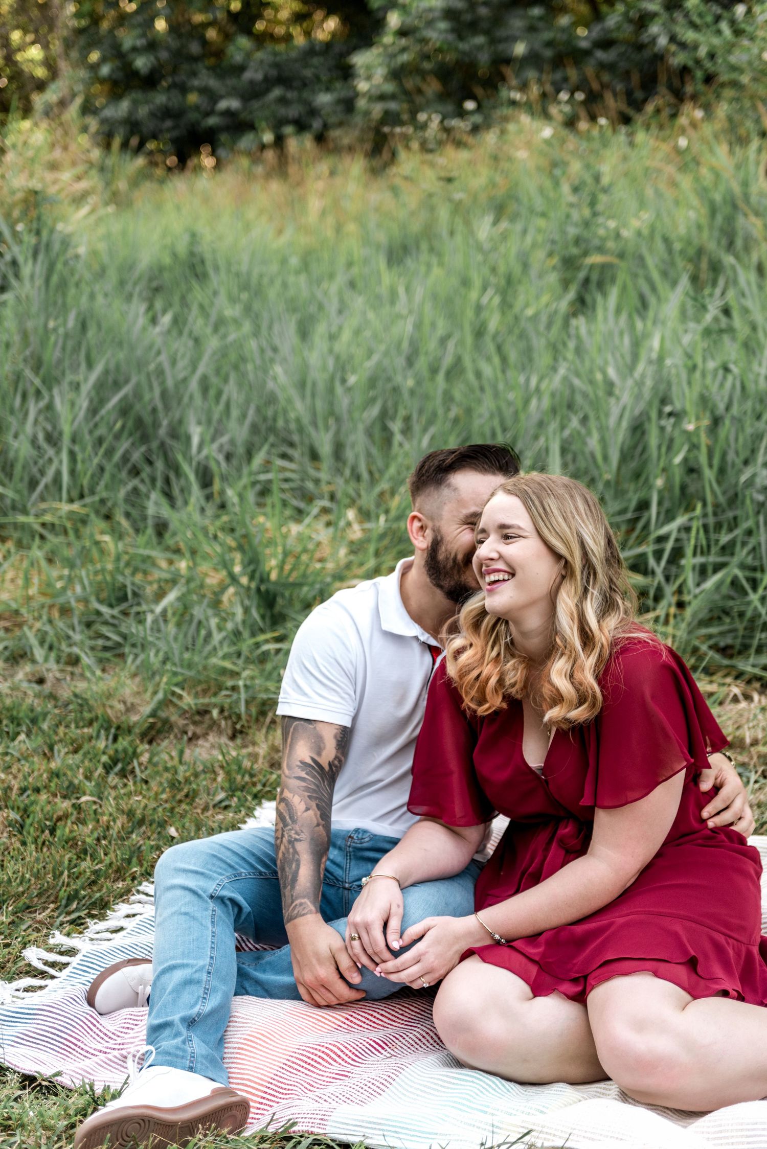 Laughing couple cuddling on a striped blanket in a lush green meadow during a sunny outdoor photo session.