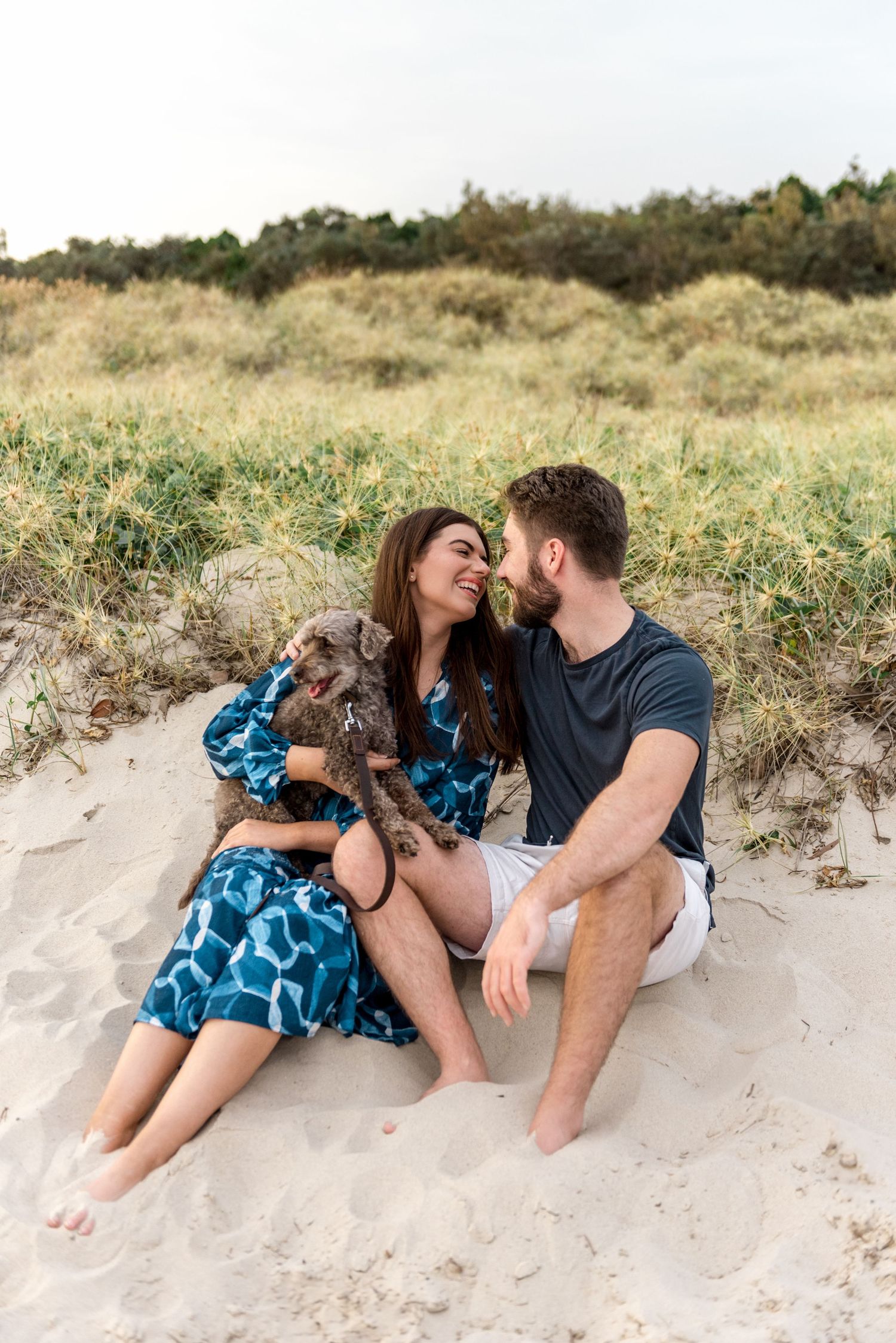 Couple sitting on beach sand dunes smiling at each other while holding their small grey dog.
