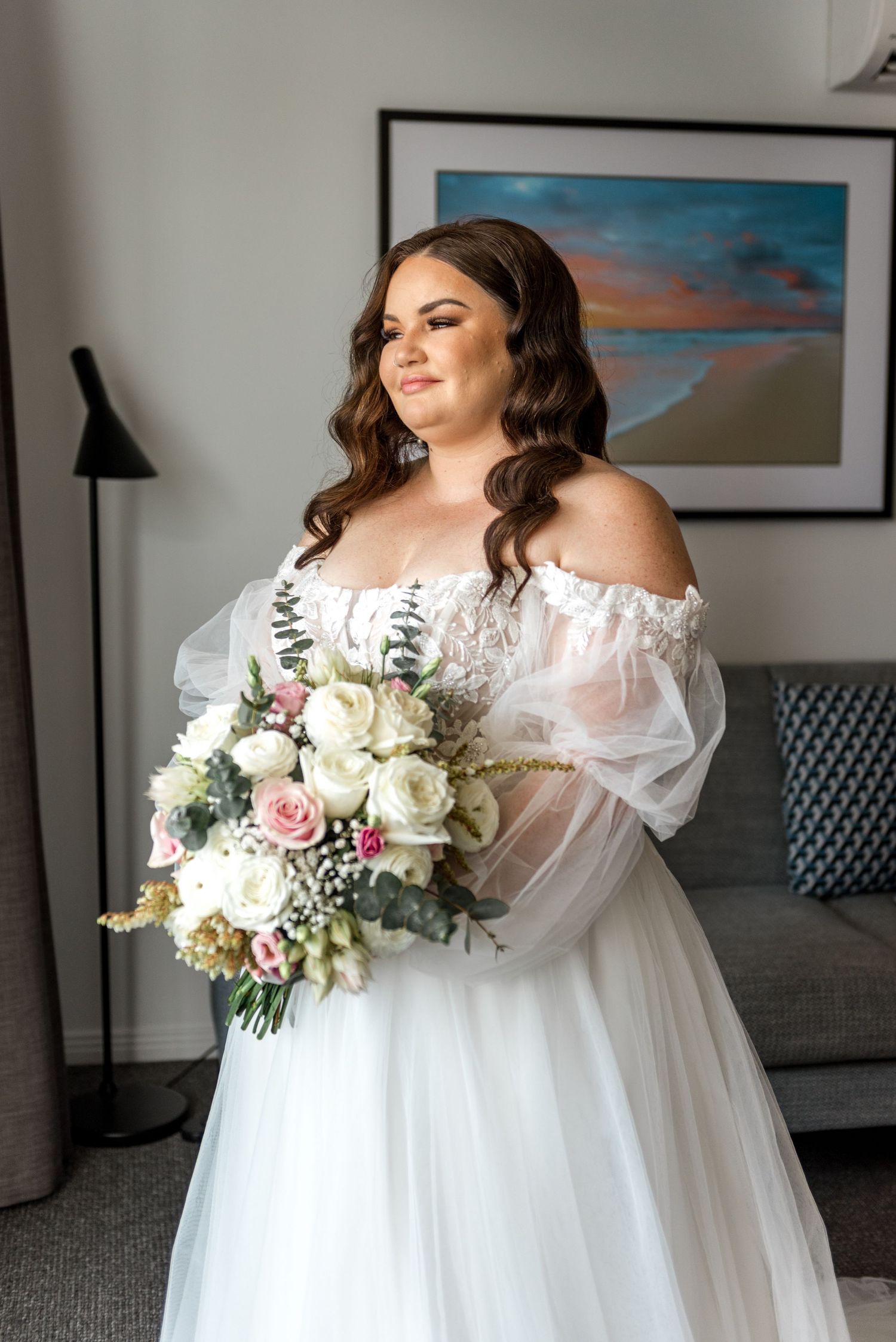 Bride in off-shoulder lace tulle gown holding pink and white rose bouquet indoors.