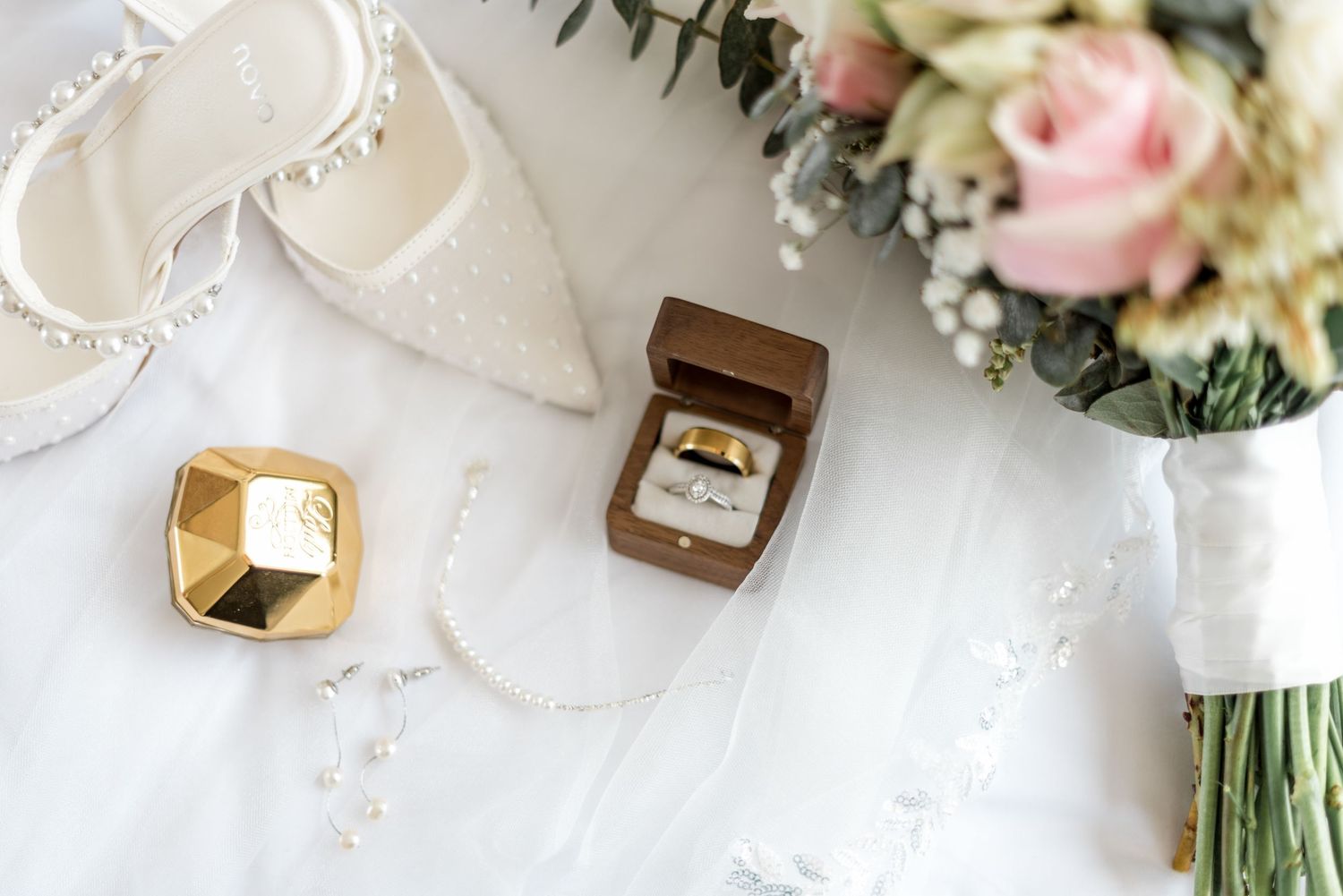 Wedding flat lay with pearl heels, gold rings in wooden box, pink bouquet, and lace veil.