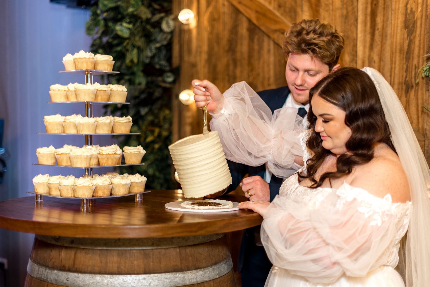 Bride and groom smile while cutting their white wedding cake surrounded by a tiered cupcake display.
