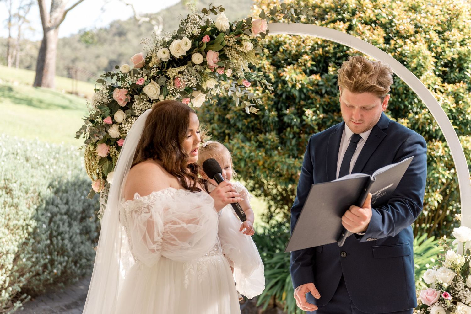 Bride reads vows at outdoor wedding ceremony beside a lush circular floral arch and officiant.