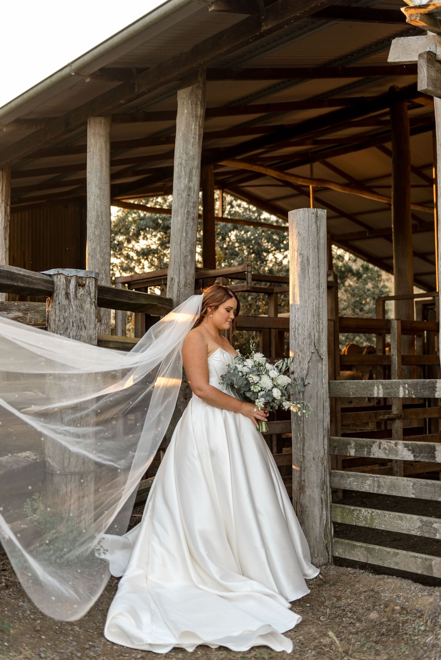 Bride in white ballgown with flowing veil beside rustic wooden barn posts holding white floral bouquet.