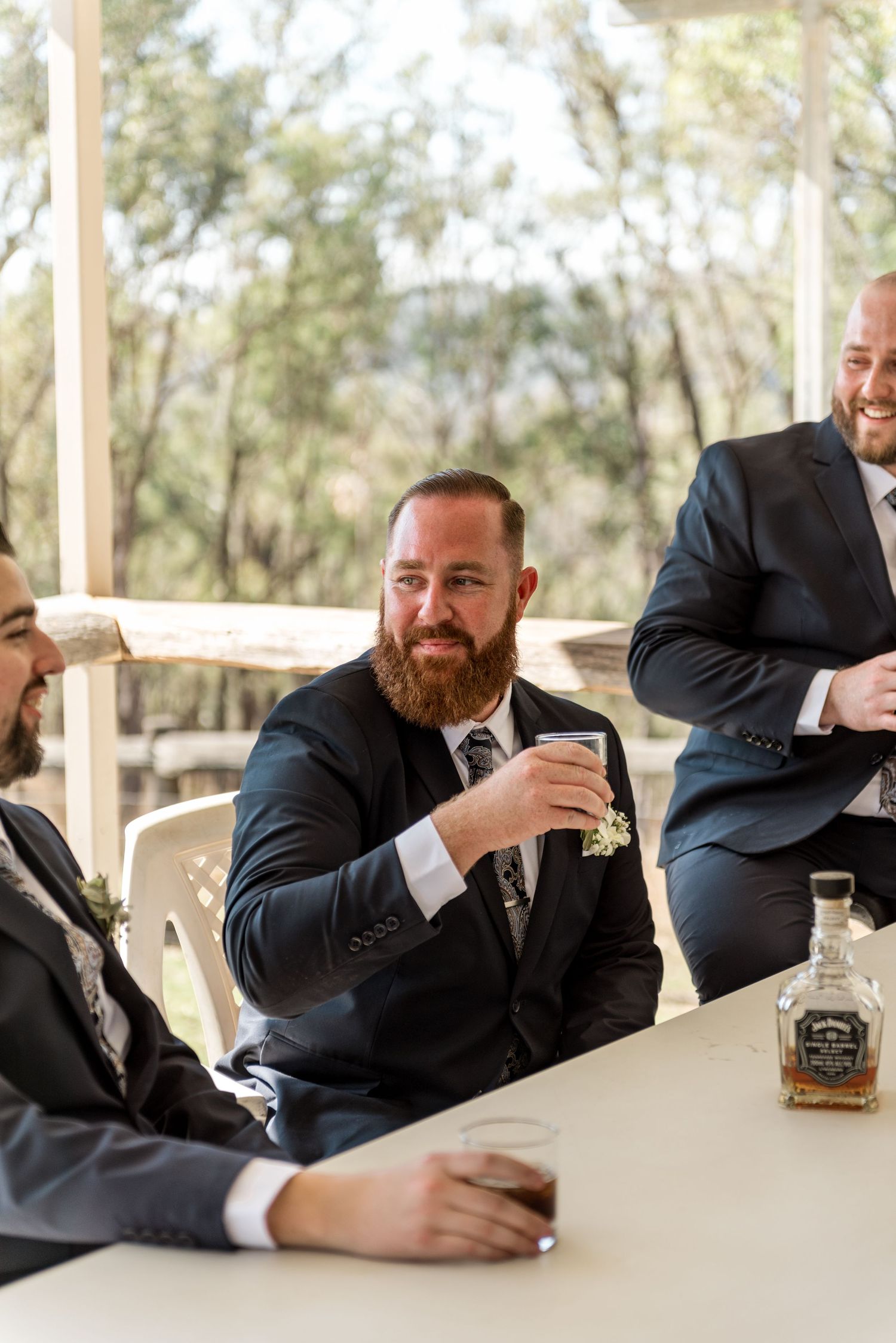 Groom with red beard toasting whiskey with groomsmen at outdoor wedding reception table.