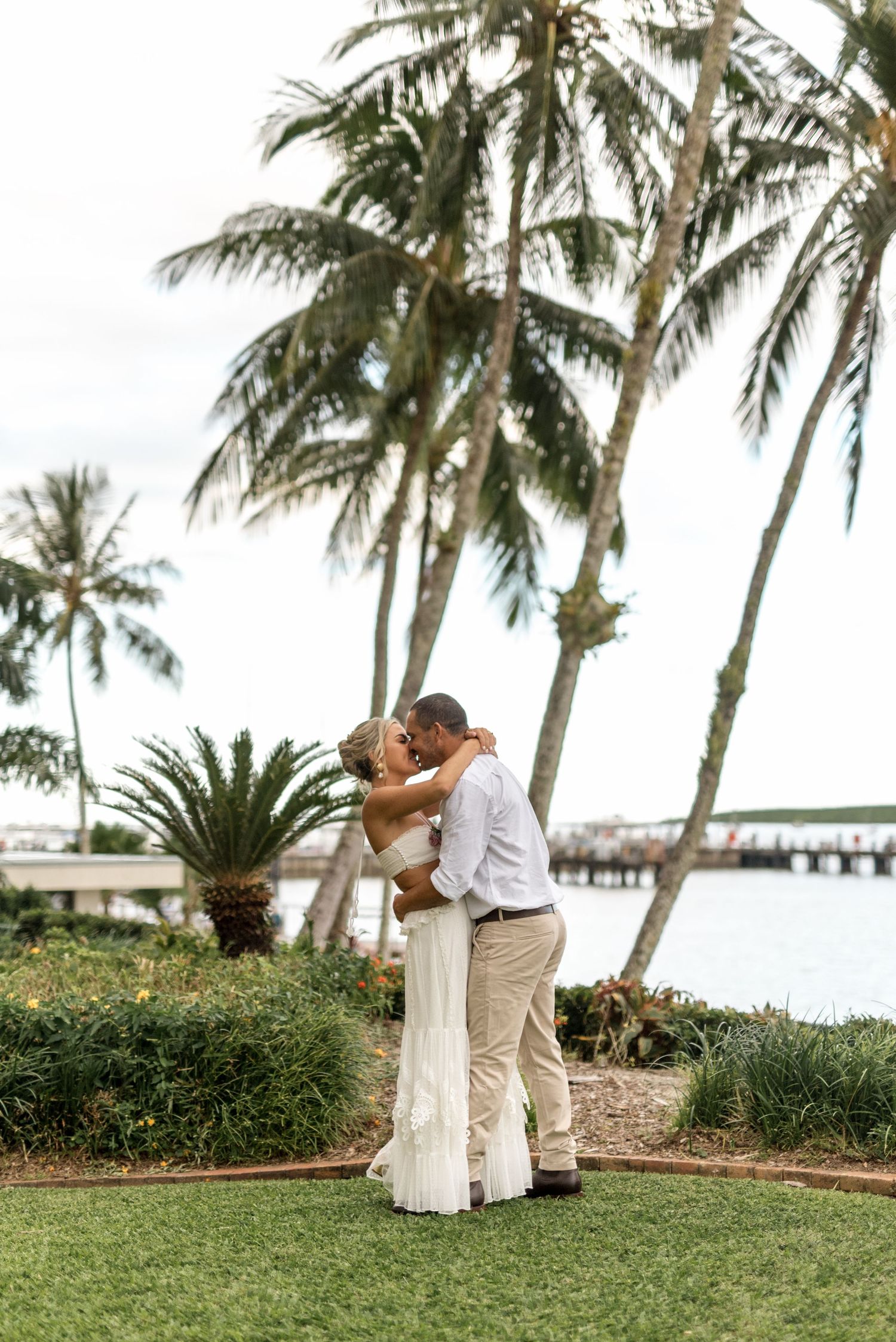 Couple shares romantic kiss surrounded by tropical palm trees with ocean and pier in background.